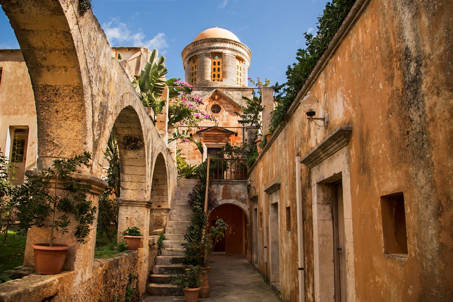 Mediterranean courtyard with stone archways, stairs and a domed tower in warm terracotta tones.