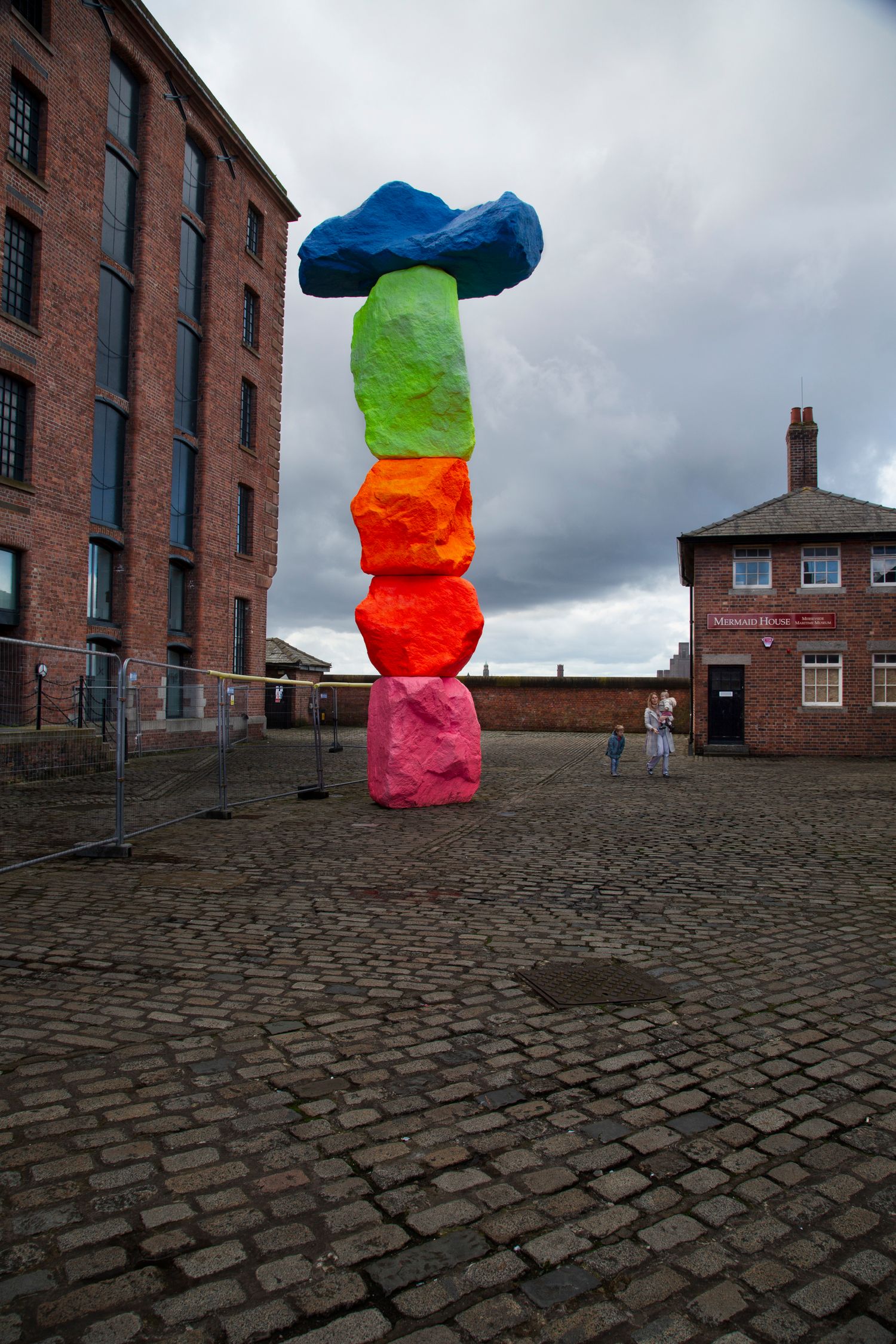 Colorful stacked art installation on cobblestone plaza in front of brick industrial building.