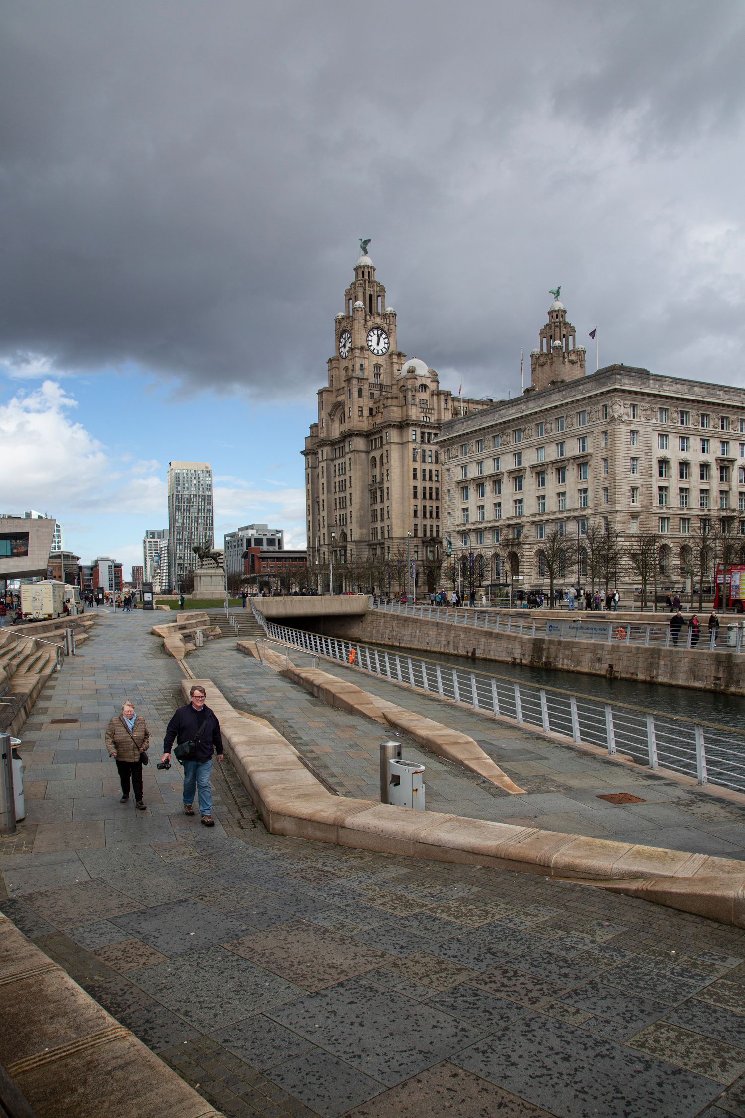 Historic waterfront buildings in Liverpool with the Liver Building visible along a riverside walkway.