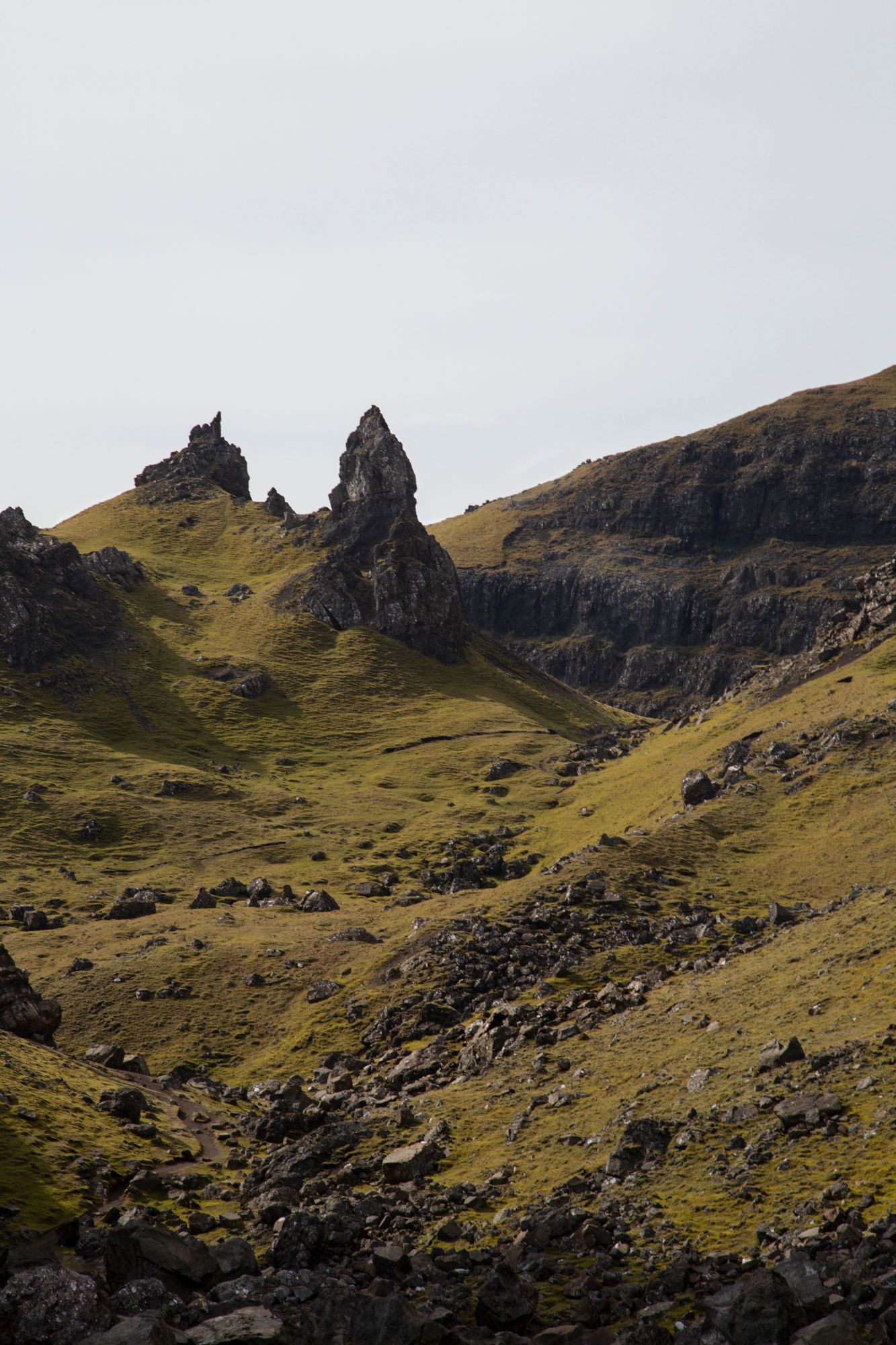 Dramatic rocky peaks rise from grassy slopes in a mountainous landscape under an overcast sky.