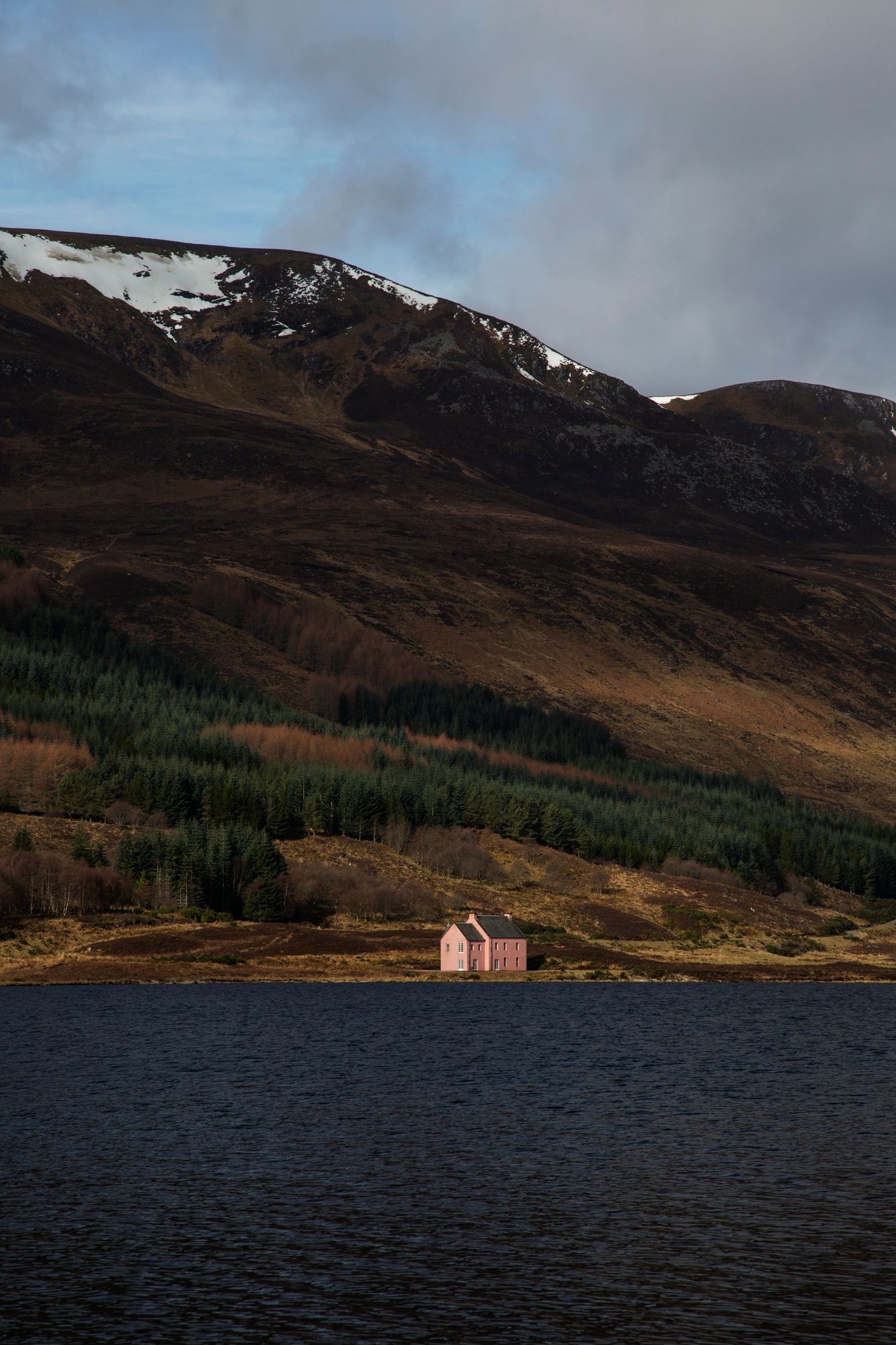 Remote pink cottage stands alone on the shores of a dark Highland loch with snow-capped mountains in the background.