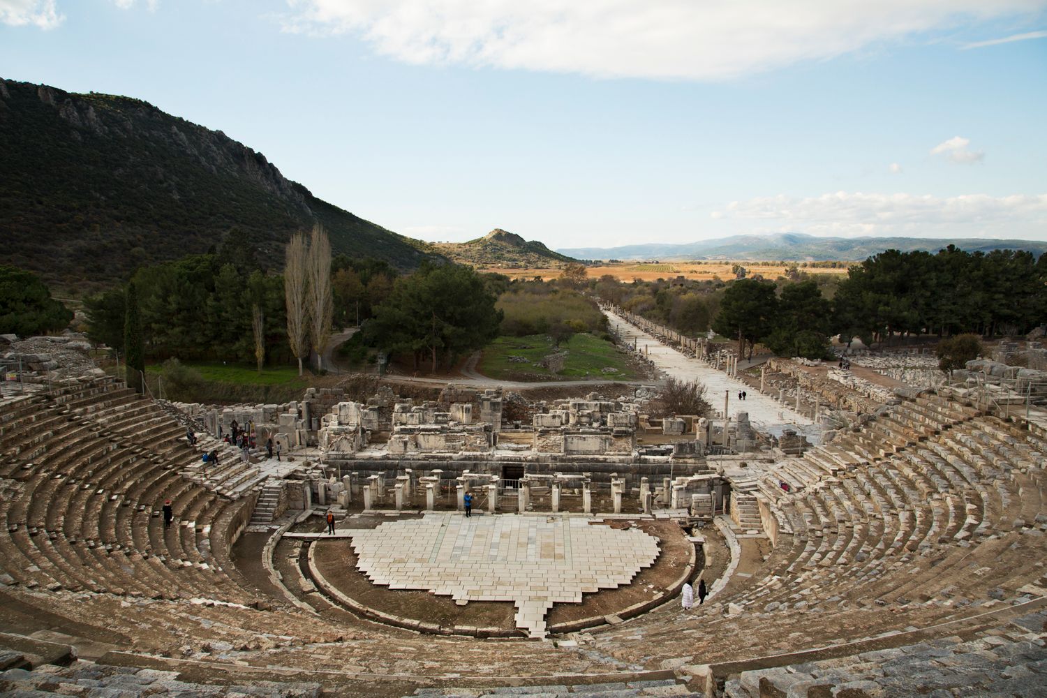 Ancient Roman amphitheater ruins with stone seating and mountain backdrop in panoramic view.