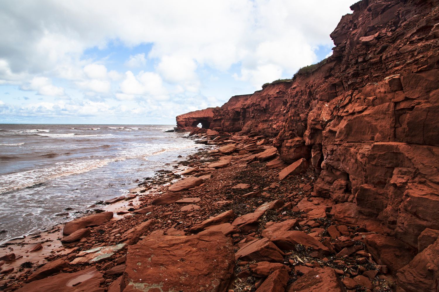 Rugged red coastal cliffs meet the ocean waves under partly cloudy skies.