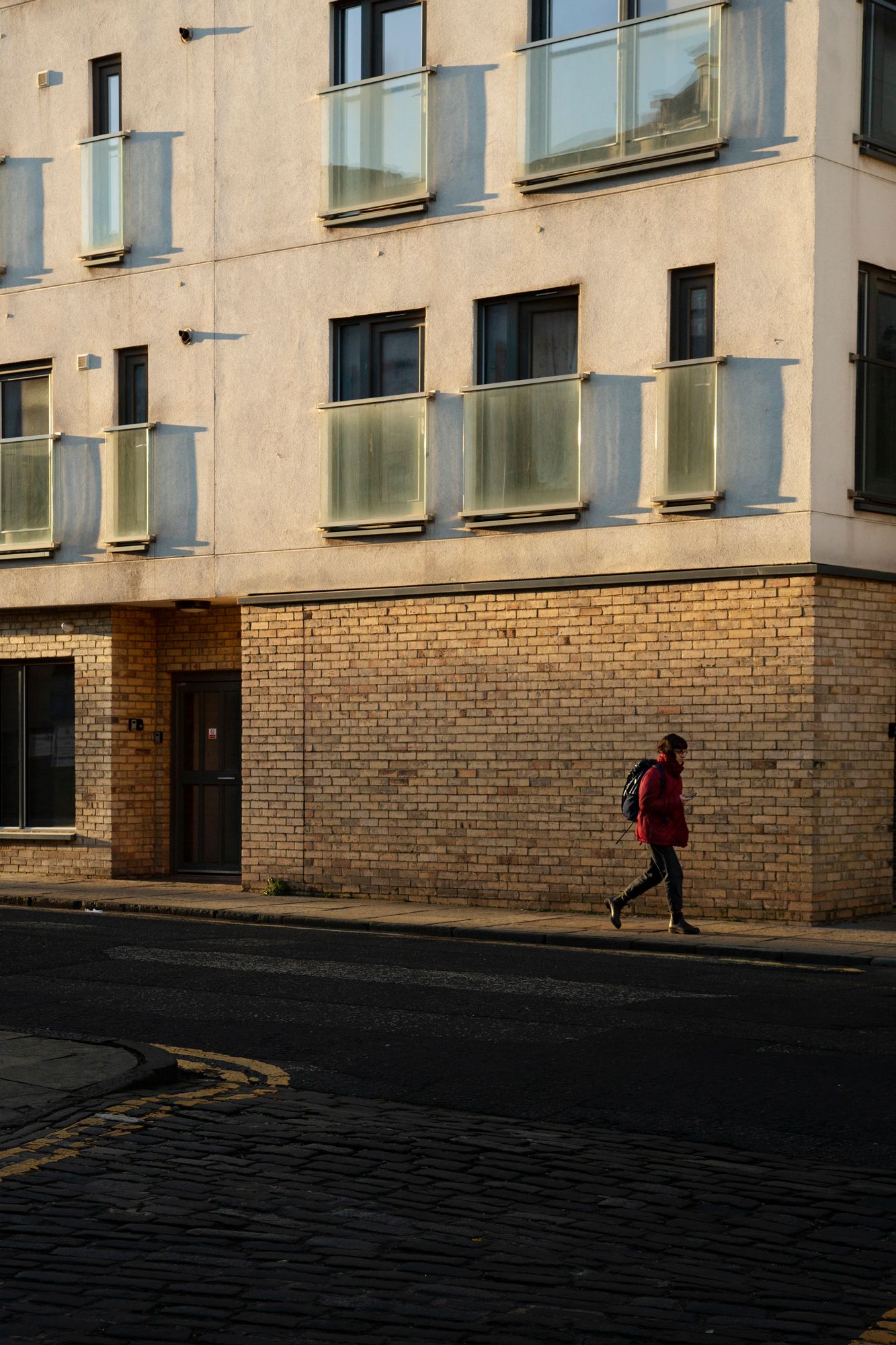 Person walking past a brick and concrete building facade with large windows casting long shadows in late afternoon light.