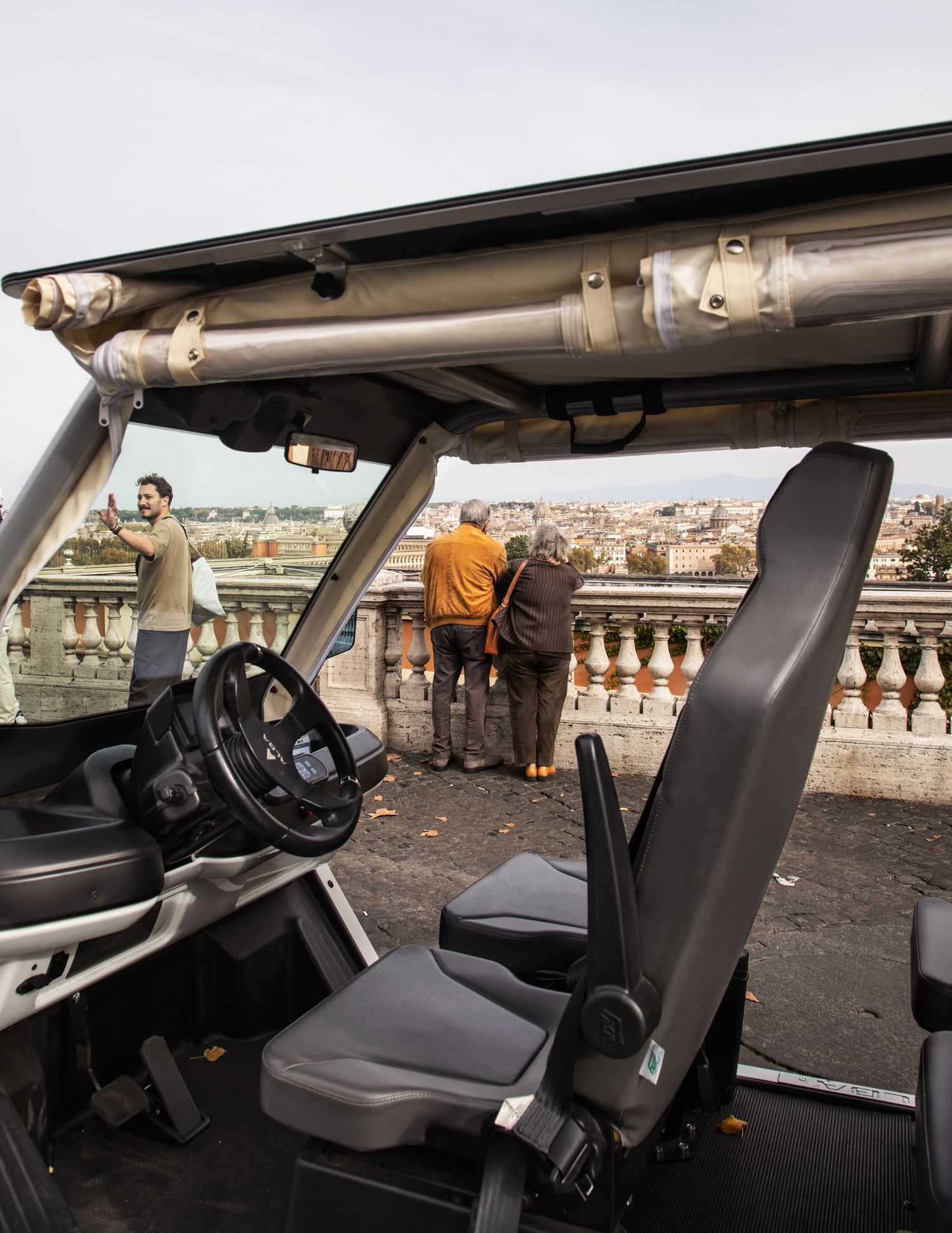 Interior view of an open-top vehicle overlooking a scenic Rome cityscape with tourists at a terrace railing.
