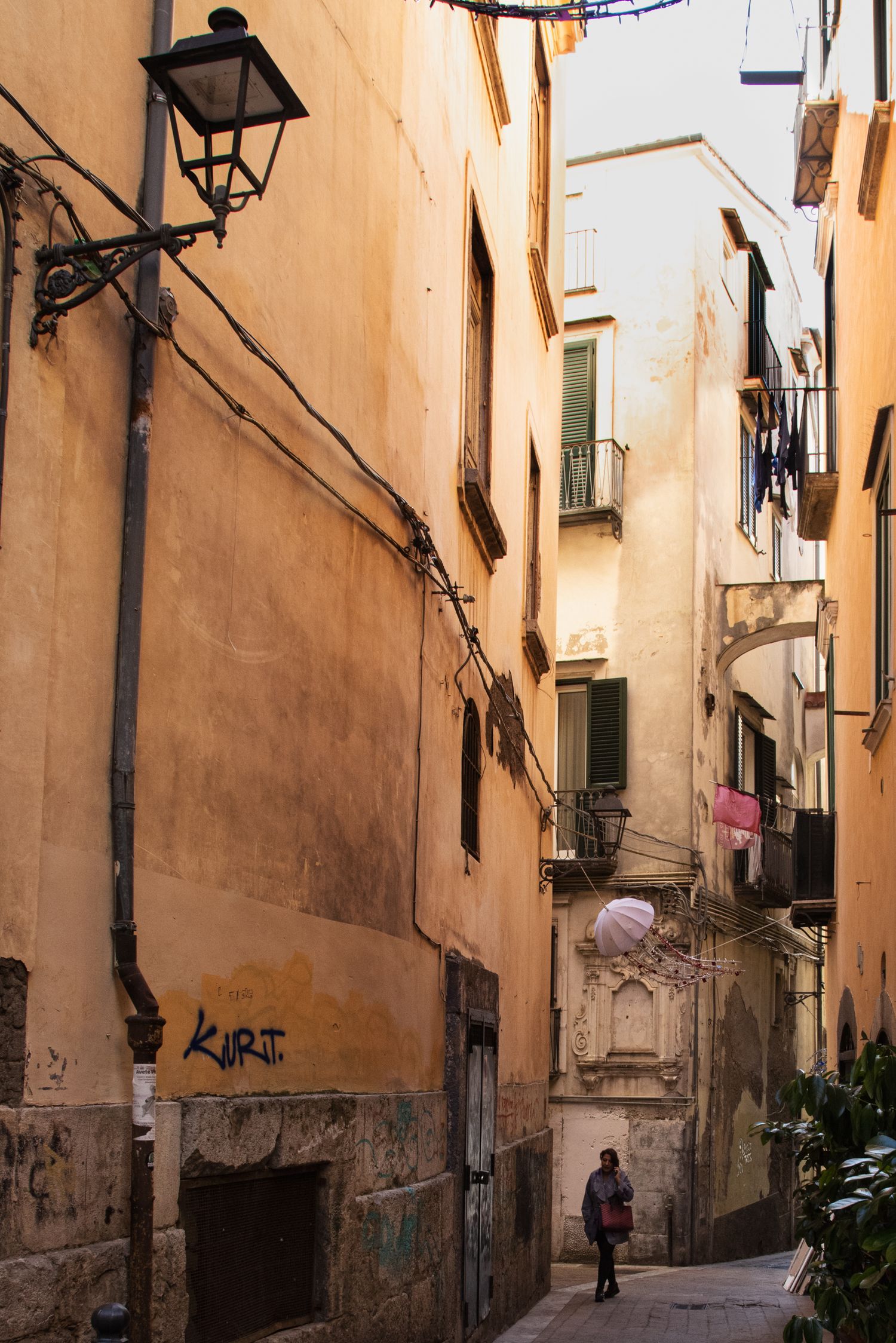 Narrow alley in an old Mediterranean city with warm orange walls, a street lamp, graffiti, and a lone pedestrian walking below.