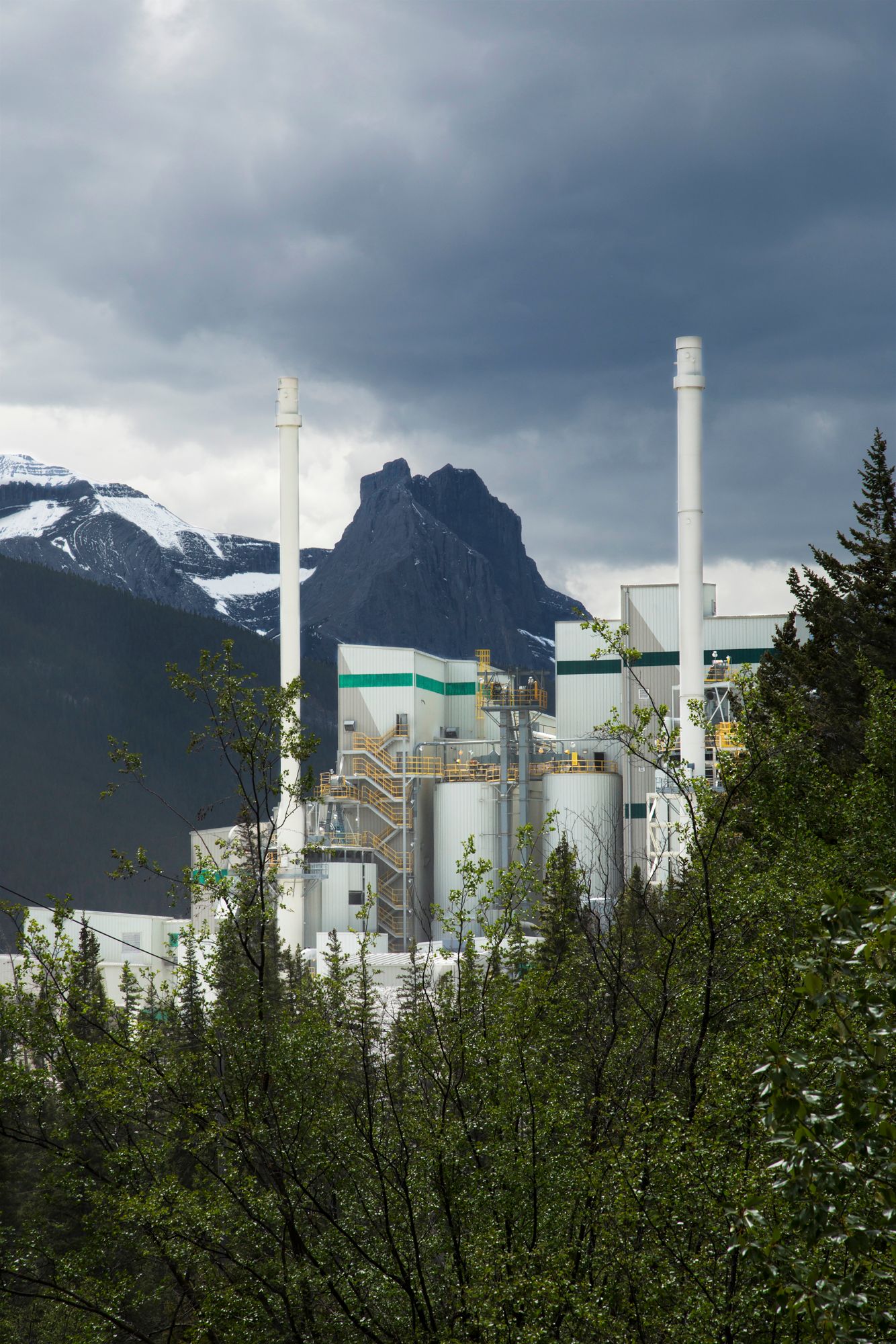 Industrial facility with white smokestacks against dramatic mountain backdrop and stormy sky.