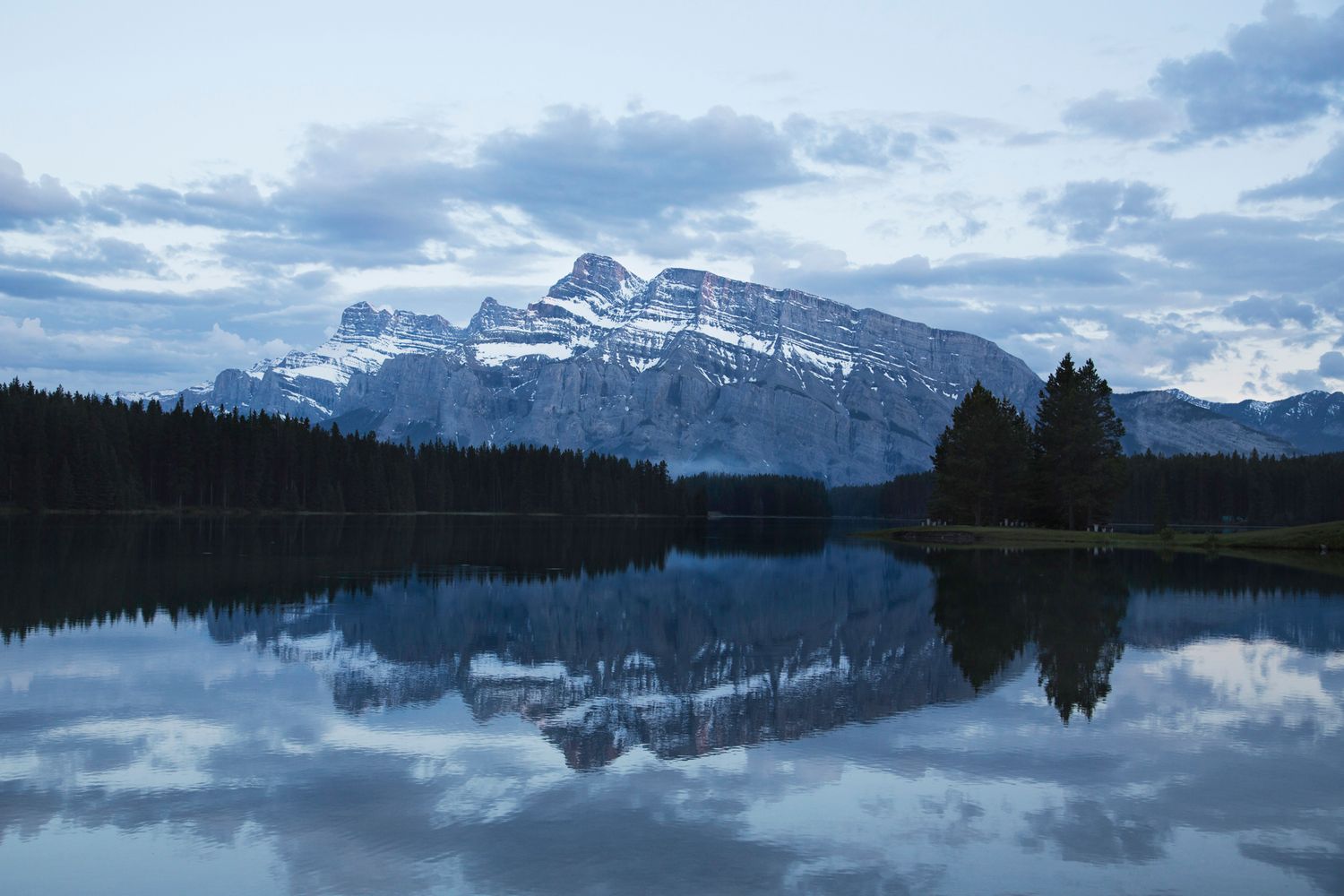Panoramic view of snow-covered mountains mirrored in clear lake waters under cloudy skies.