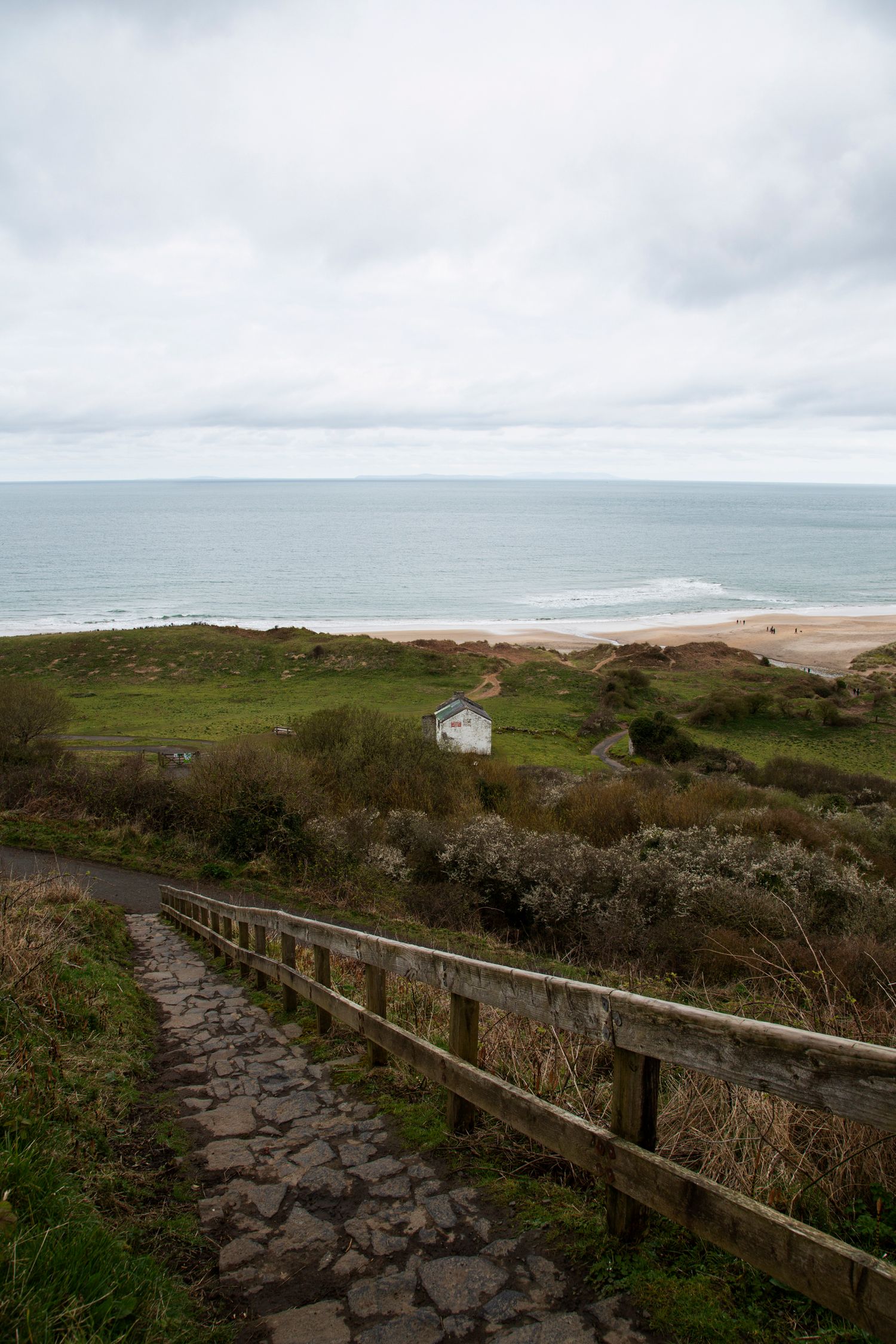 Stone pathway with wooden railing descends towards sandy beach and ocean vista.