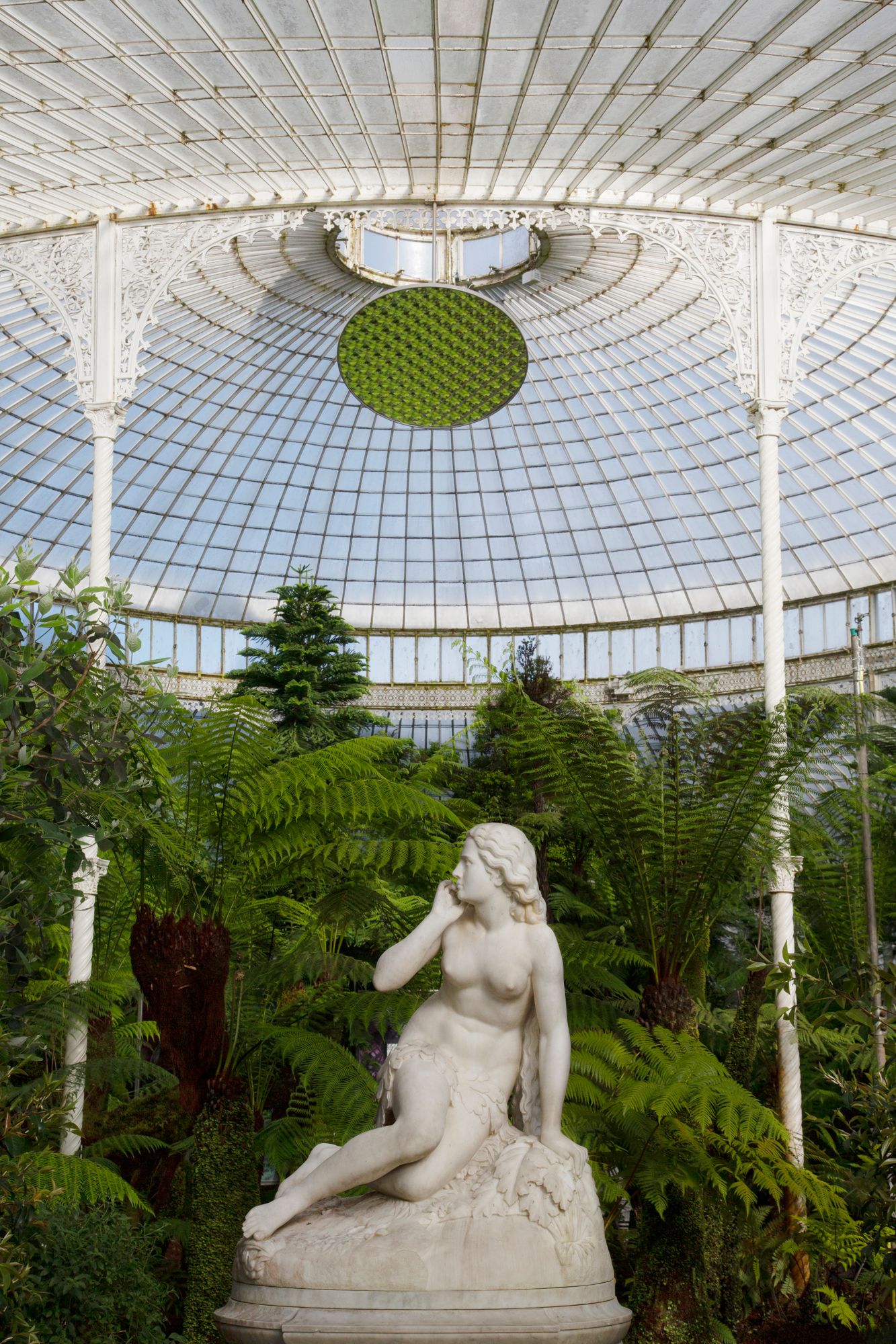 Classical marble statue surrounded by lush ferns under an ornate Victorian glass dome conservatory ceiling.