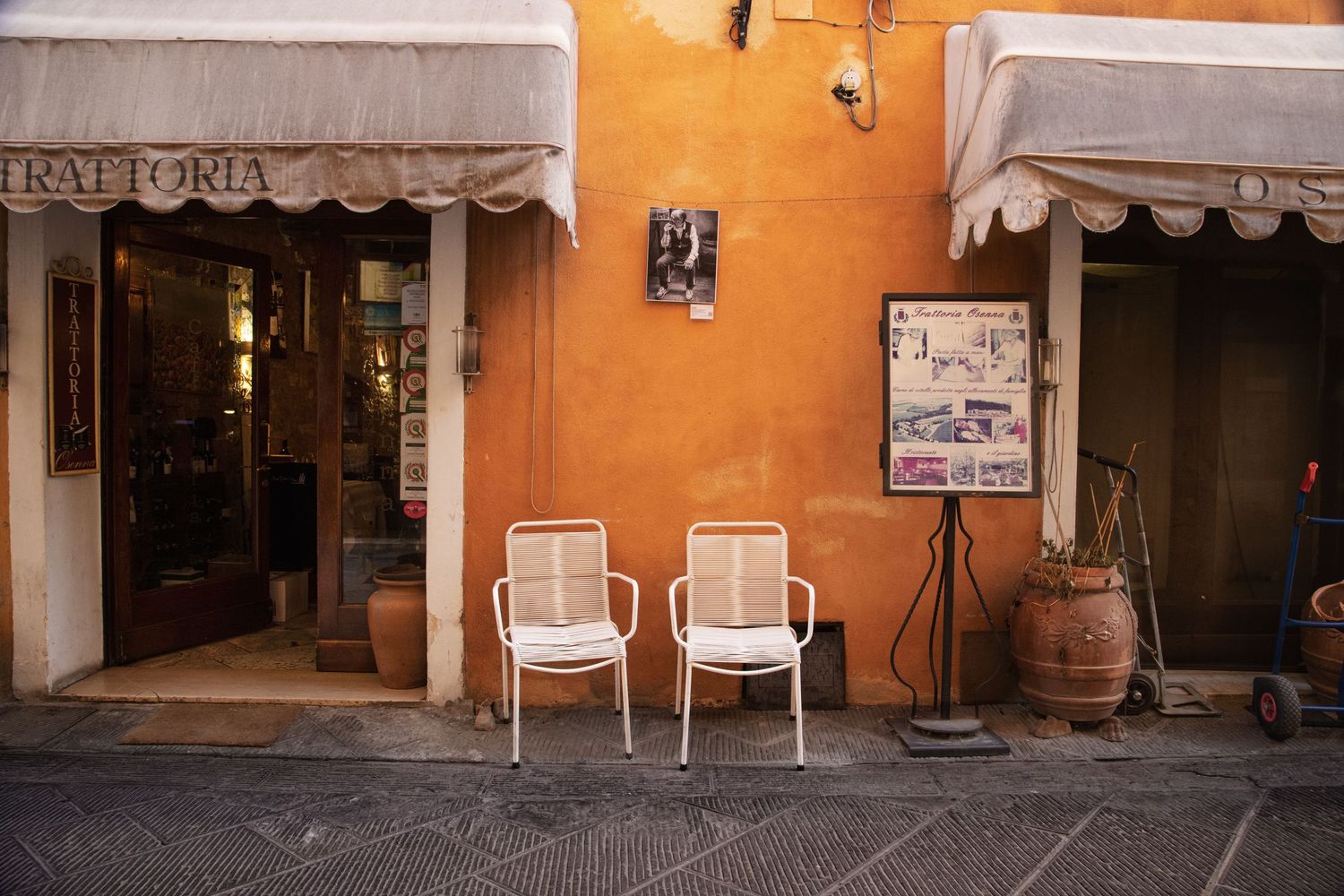 White wicker chairs outside a rustic Italian trattoria with orange walls, wooden barrel, and menu board on cobblestone street.