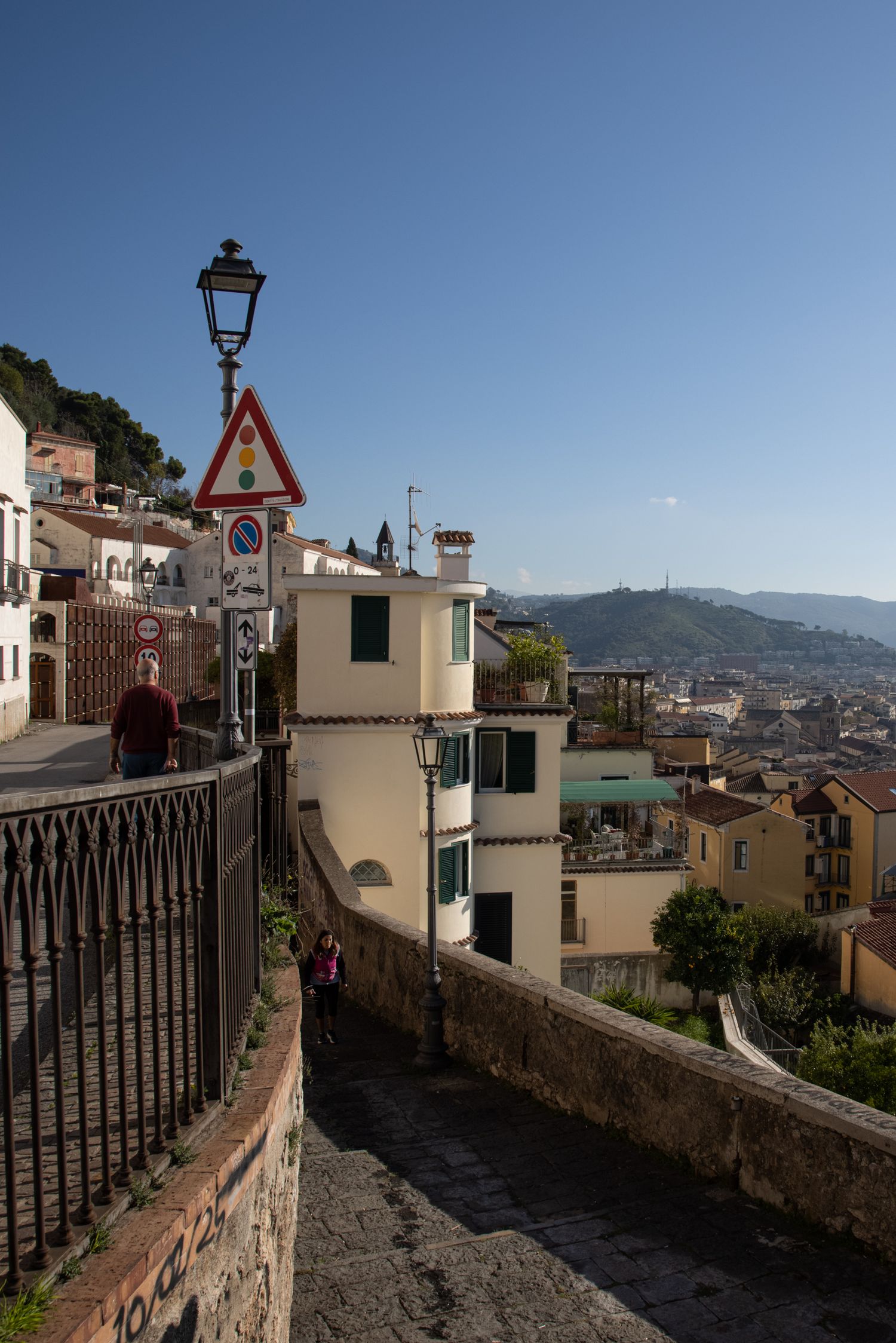 Scenic Italian hillside street with iron railing, street lamp, road signs, and panoramic city view under clear blue sky.