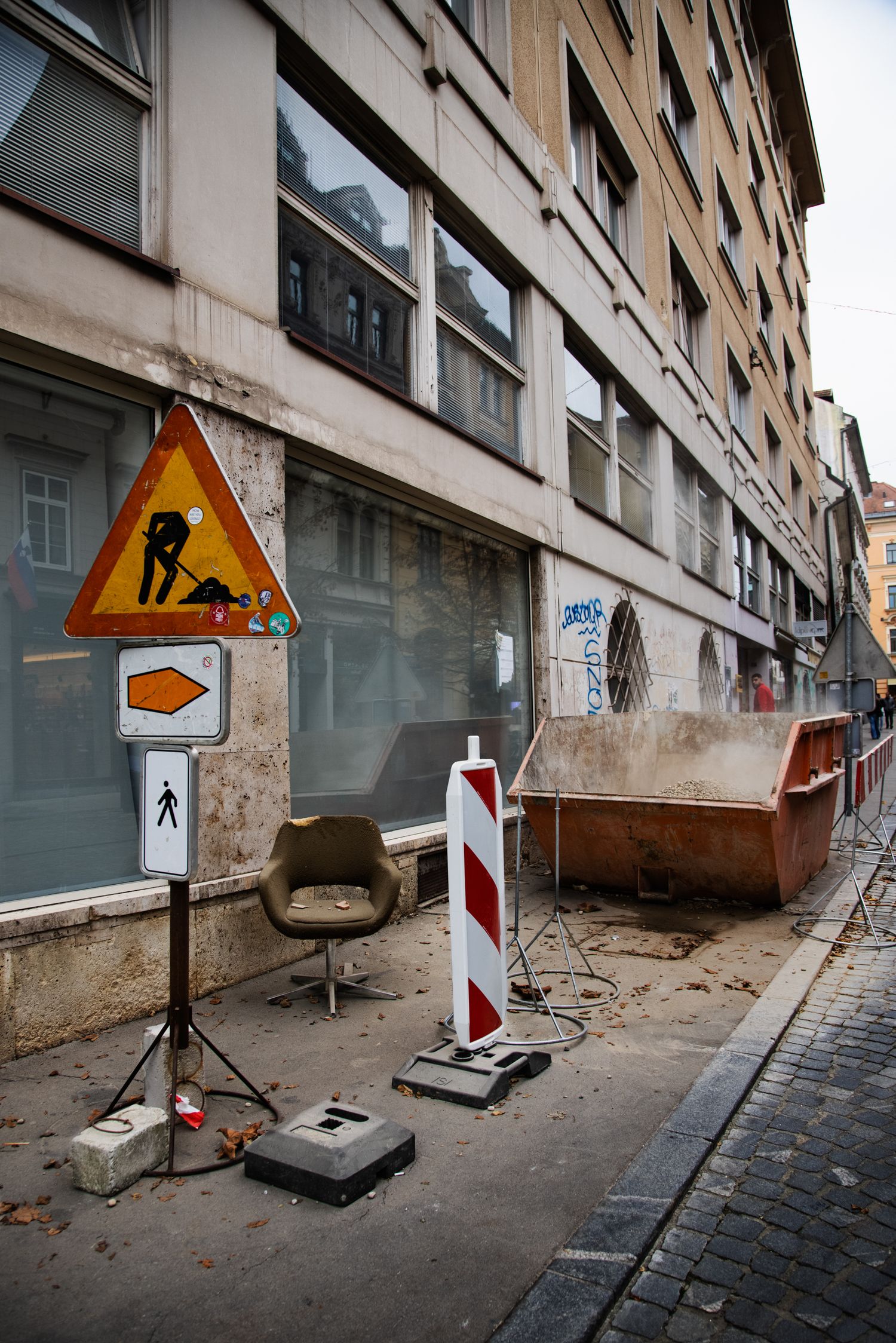 Urban street construction zone with warning signs, a chair, and a dumpster beside a weathered building facade.