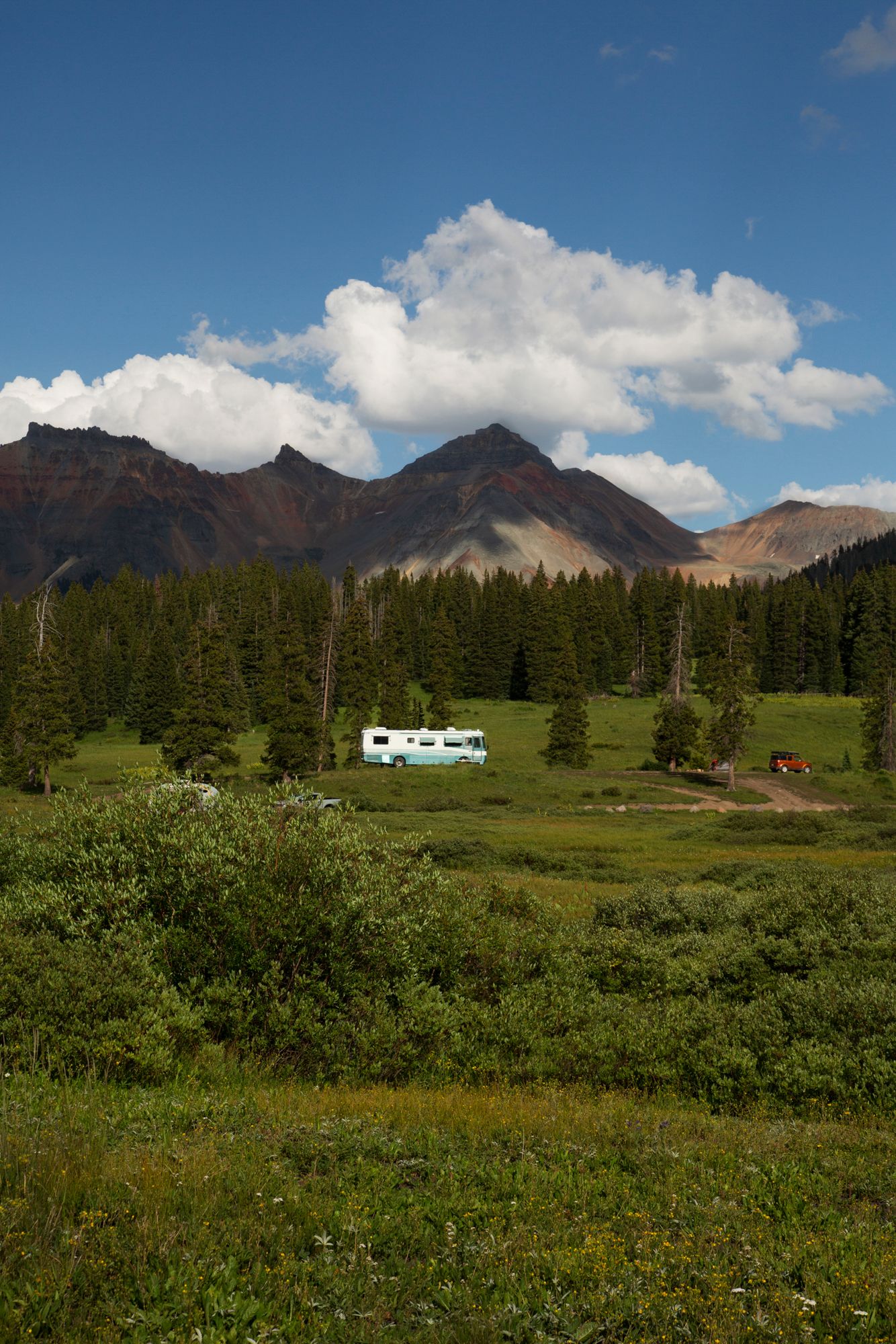 Mountain landscape with green meadows, evergreen forest, and a small cabin nestled beneath rugged peaks and puffy clouds.