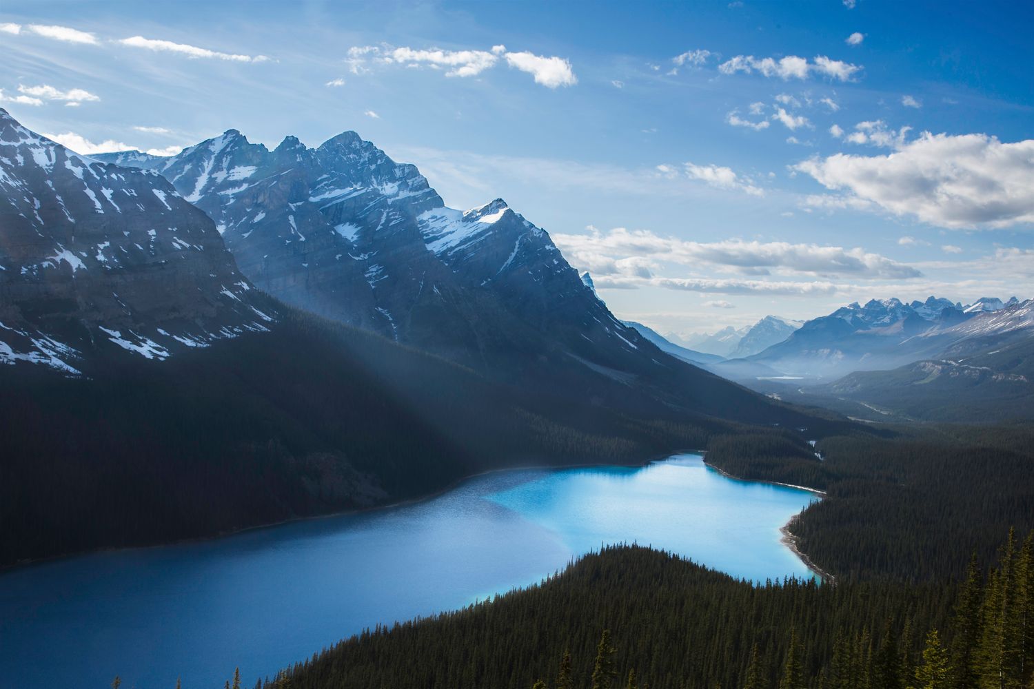 Dramatic mountain range with snow-covered peaks rises against a bright blue sky over wilderness terrain.