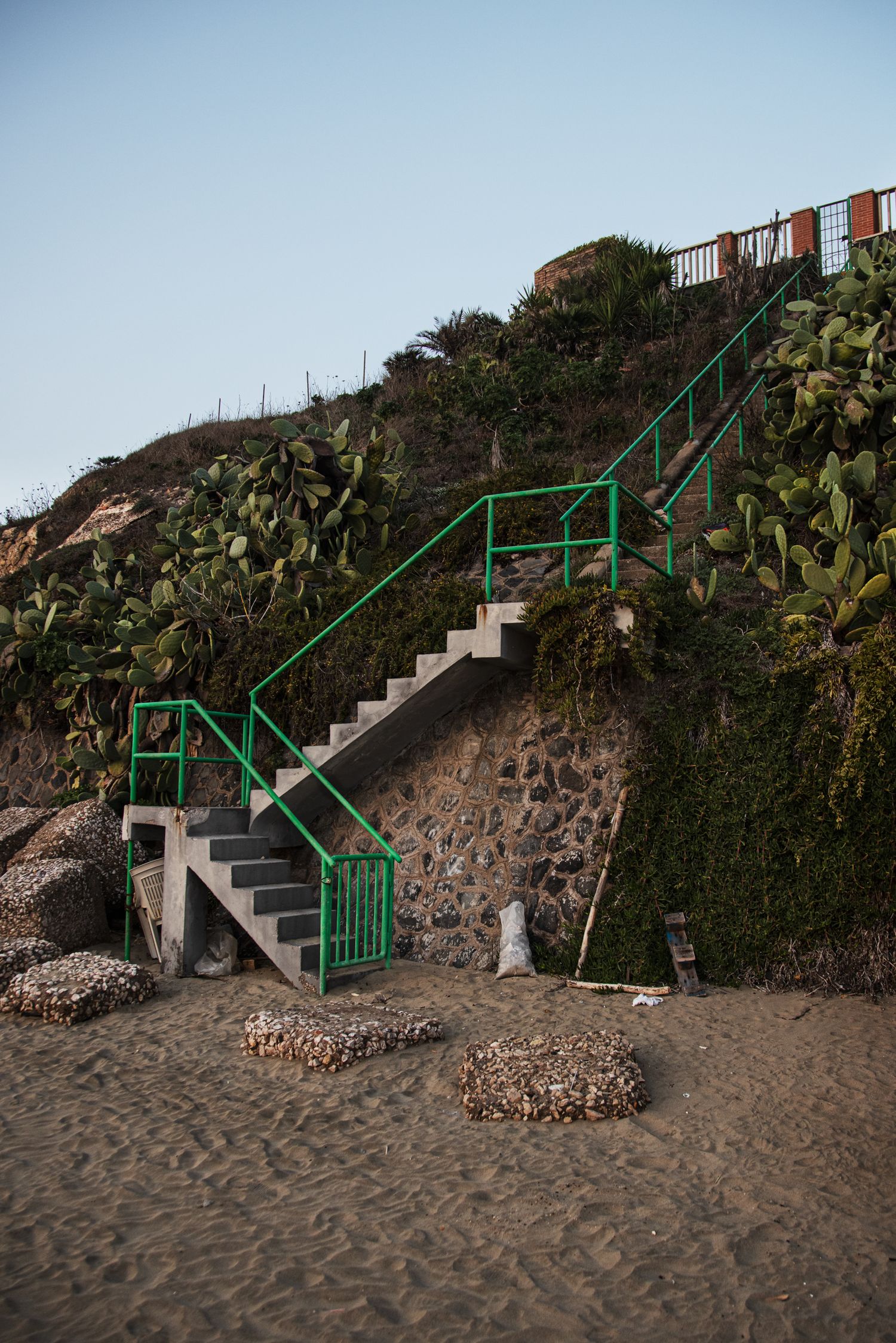 Concrete beach stairs with green railings lead up a rocky hillside with cacti and lush greenery under a clear blue sky.
