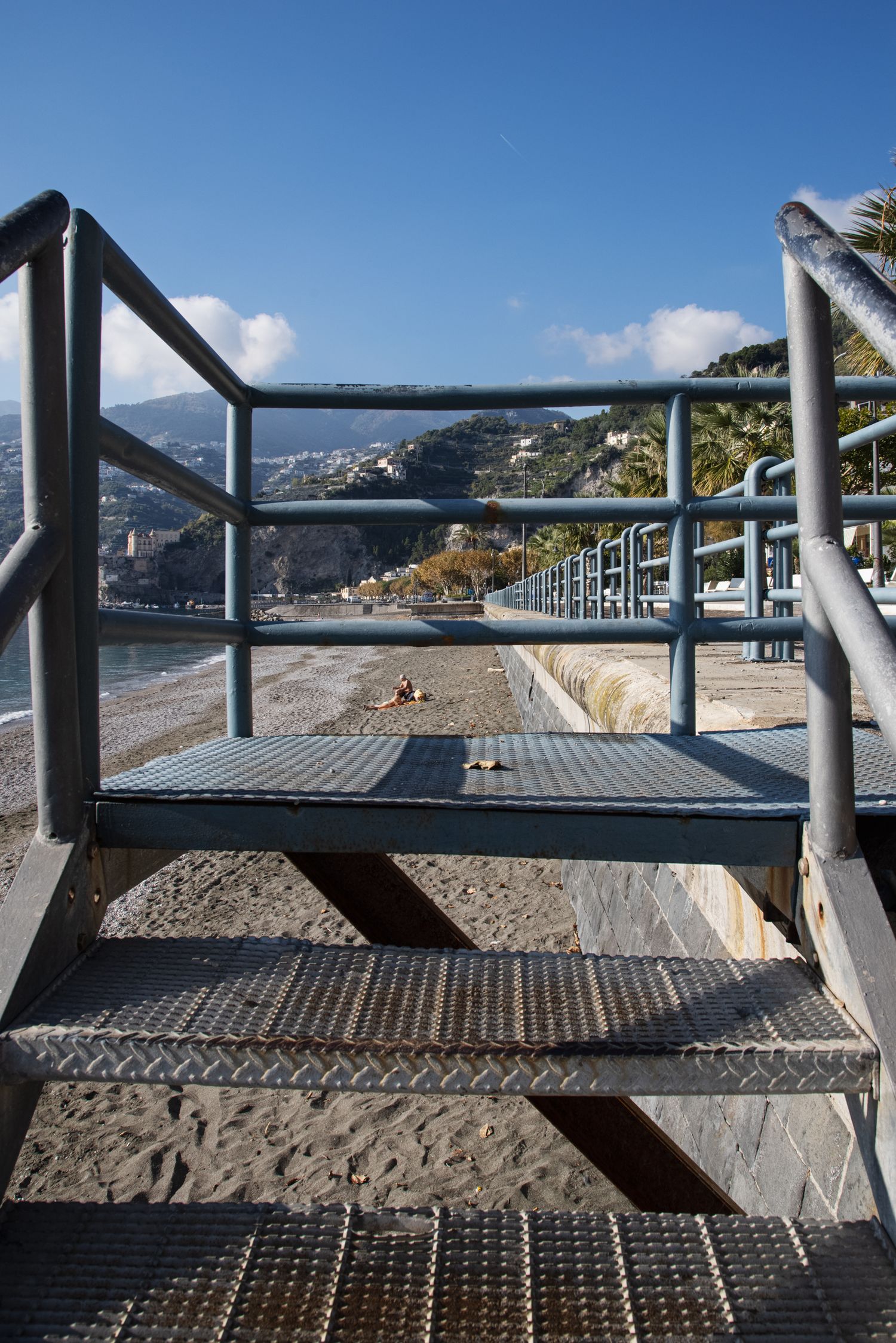 Metal staircase with diamond plate steps leads to a sandy beach with mountain views under a clear blue sky.