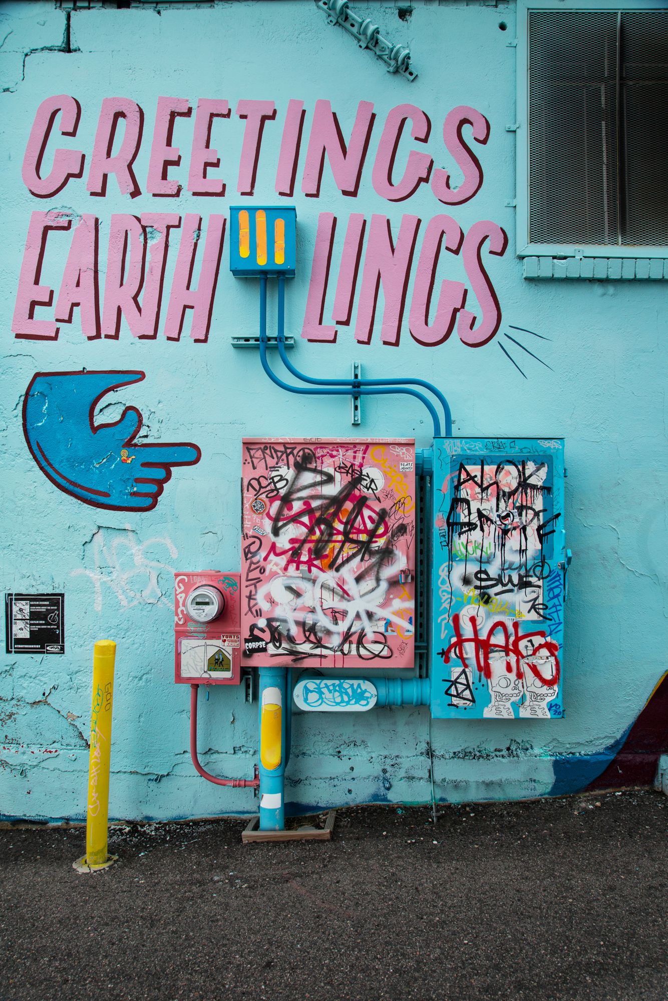 Urban street art mural with pink graffiti text reading 'GREETINGS' on turquoise wall with smaller artworks displayed below.