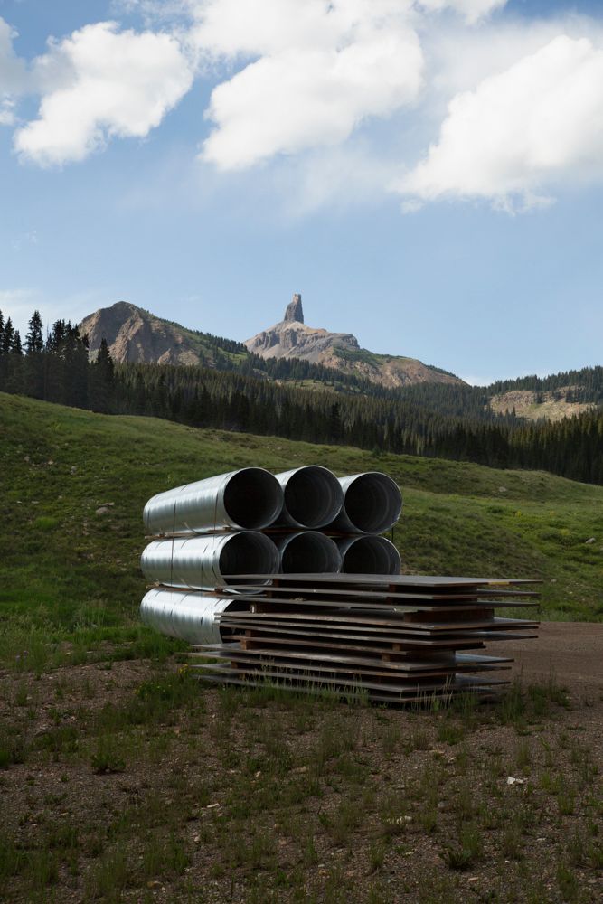 Large industrial pipes stacked on hillside with mountain peaks rising in background.