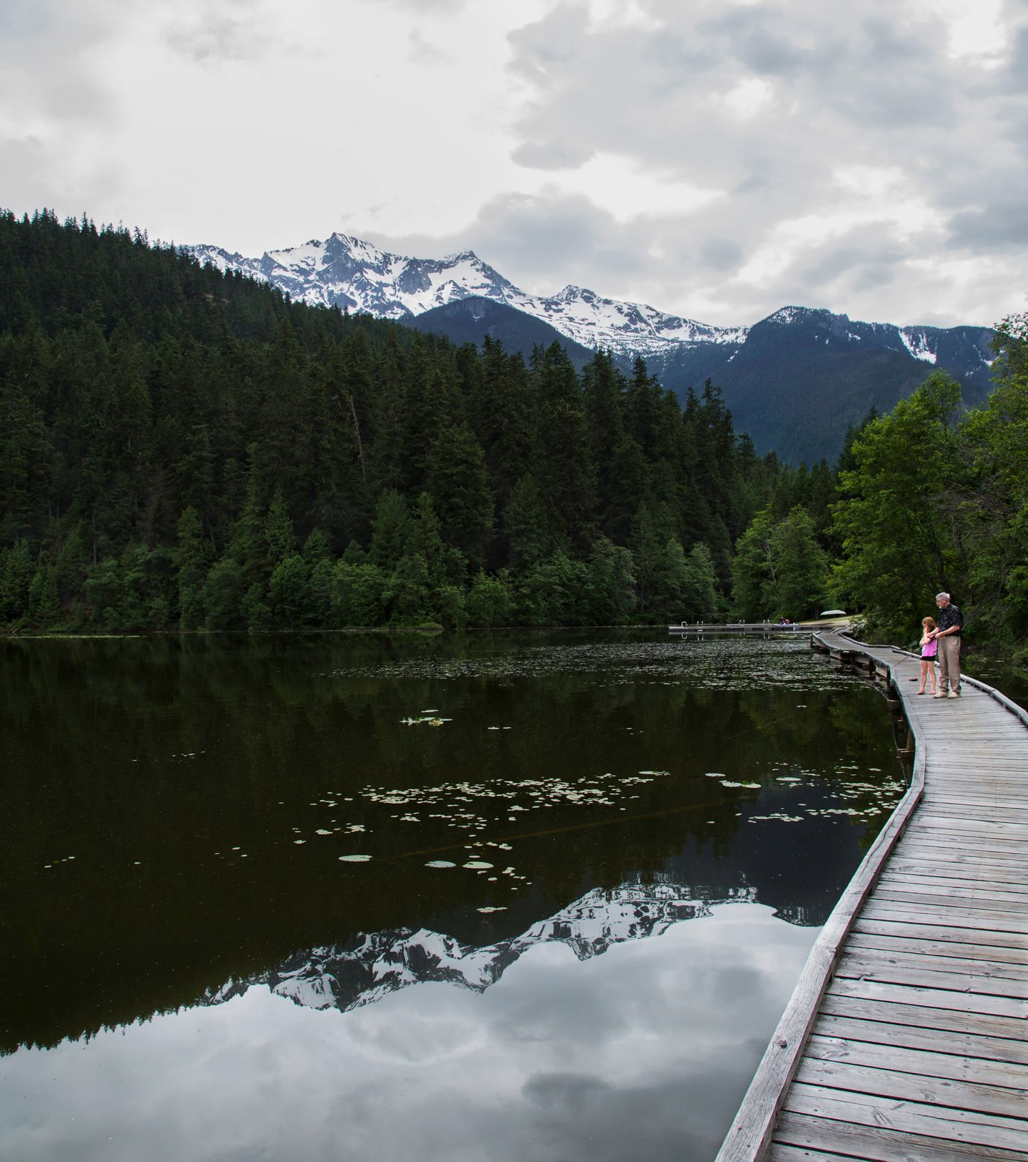 Wooden boardwalk extends over reflective mountain lake with snow-capped peaks in background.