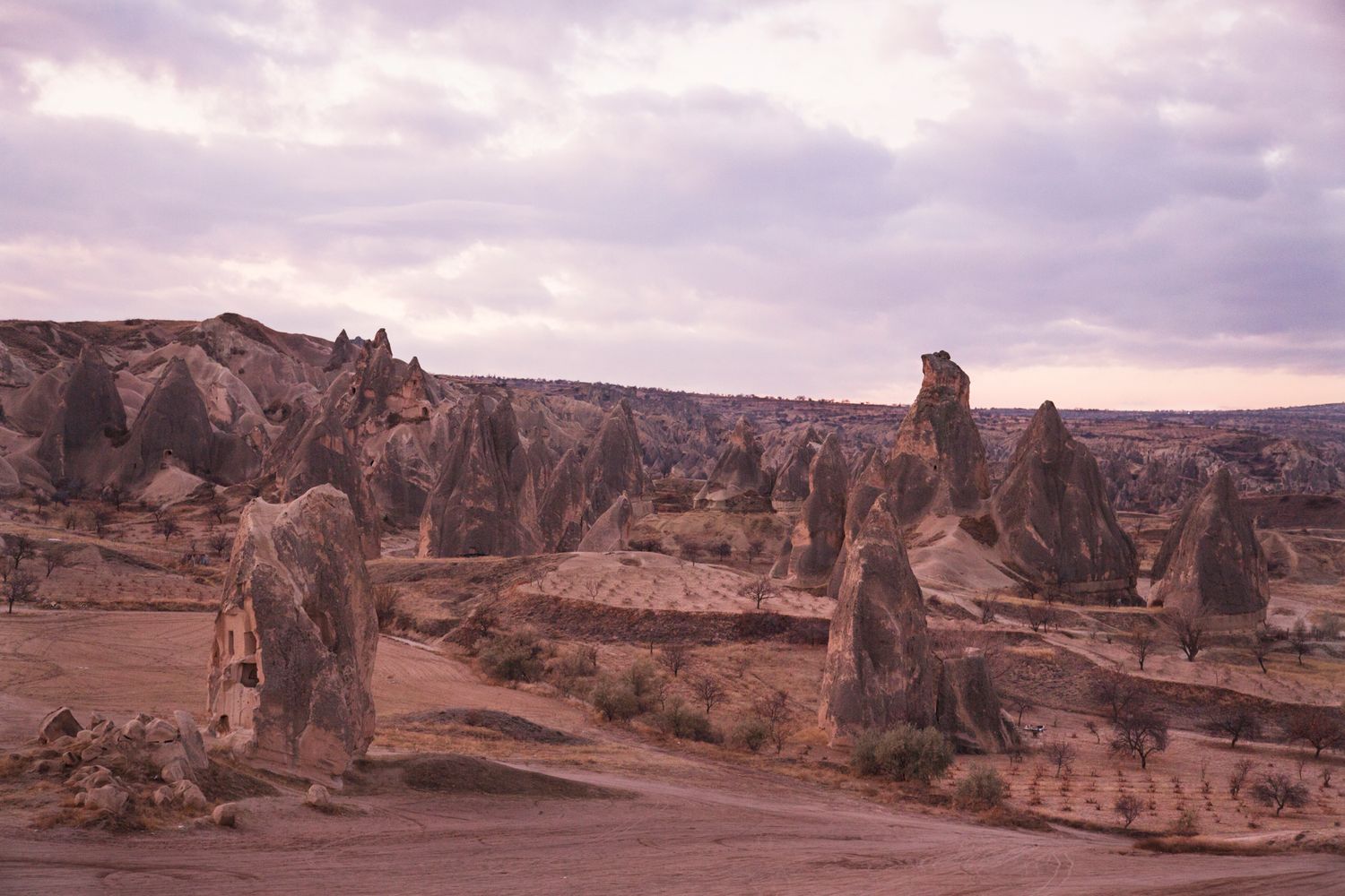 Ancient rock pillars and carved dwellings stand against a cloudy sky in a desert landscape.