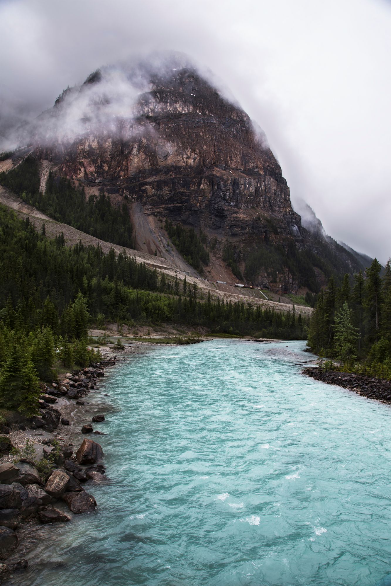 Turquoise glacial river flows past misty mountain face in Canadian Rockies.