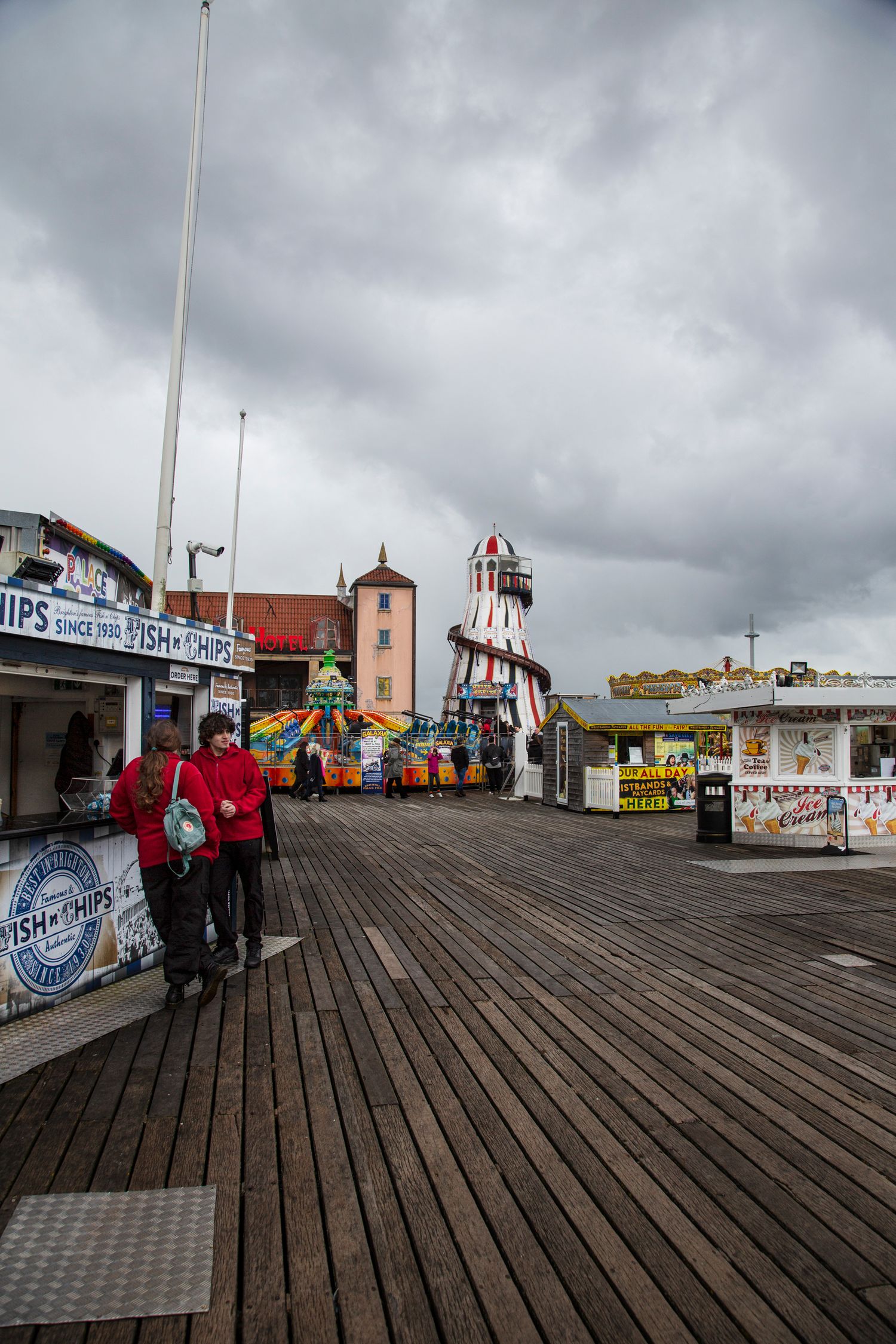 Wooden boardwalk on Brighton Pier with fairground rides and cloudy sky.