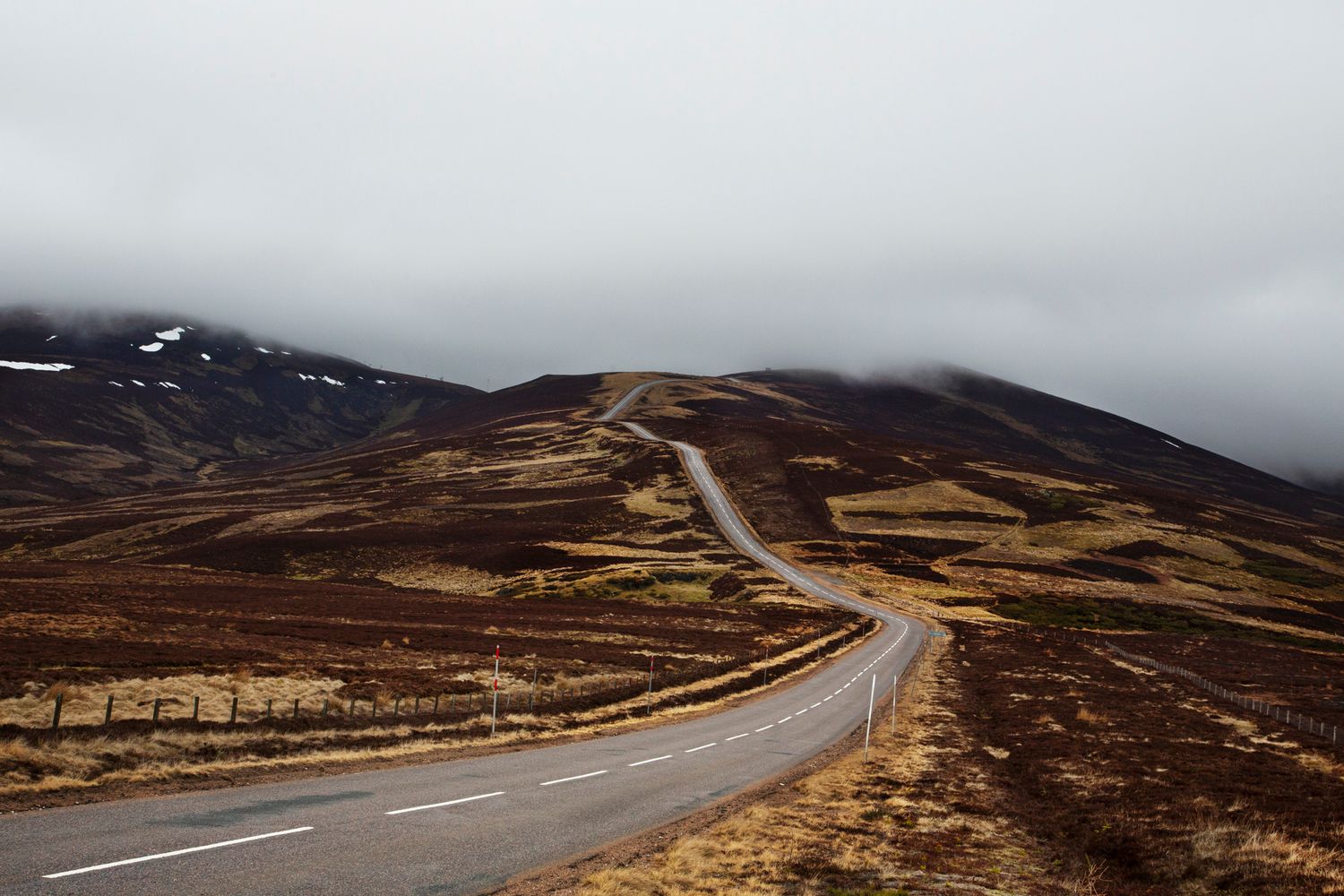 A scenic winding road cuts through the rugged Scottish Highlands with mountains and moody skies in the background.