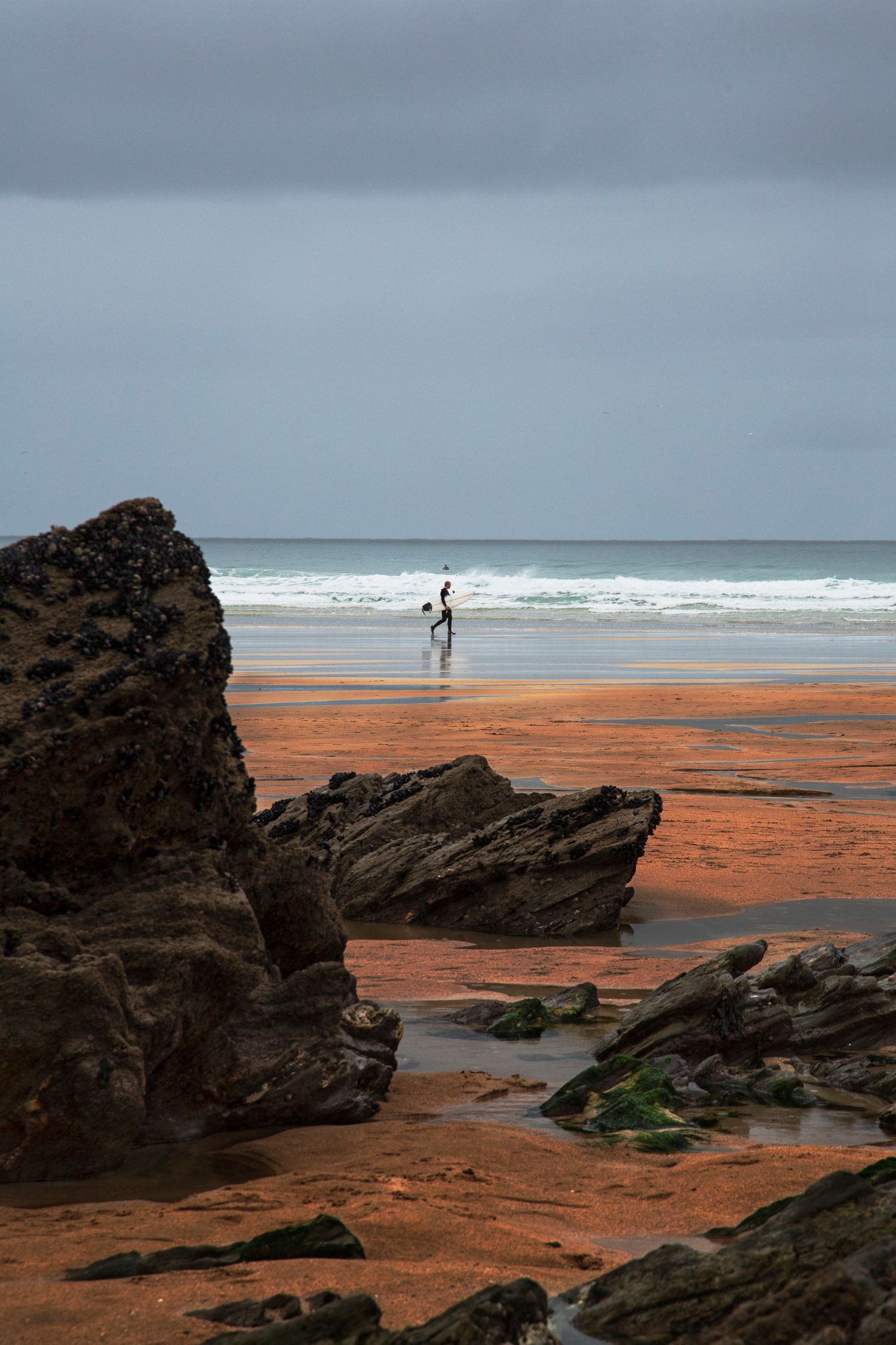 Orange sandy beach with dark rocky outcrops and distant surfers against a moody sky.