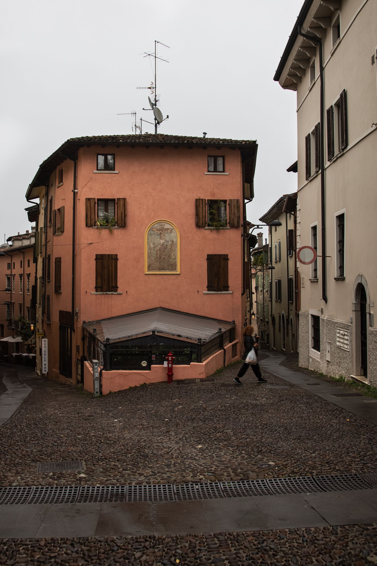 Cobblestone street corner in an Italian town featuring a terracotta building with wooden shutters and a religious fresco on a cloudy day.