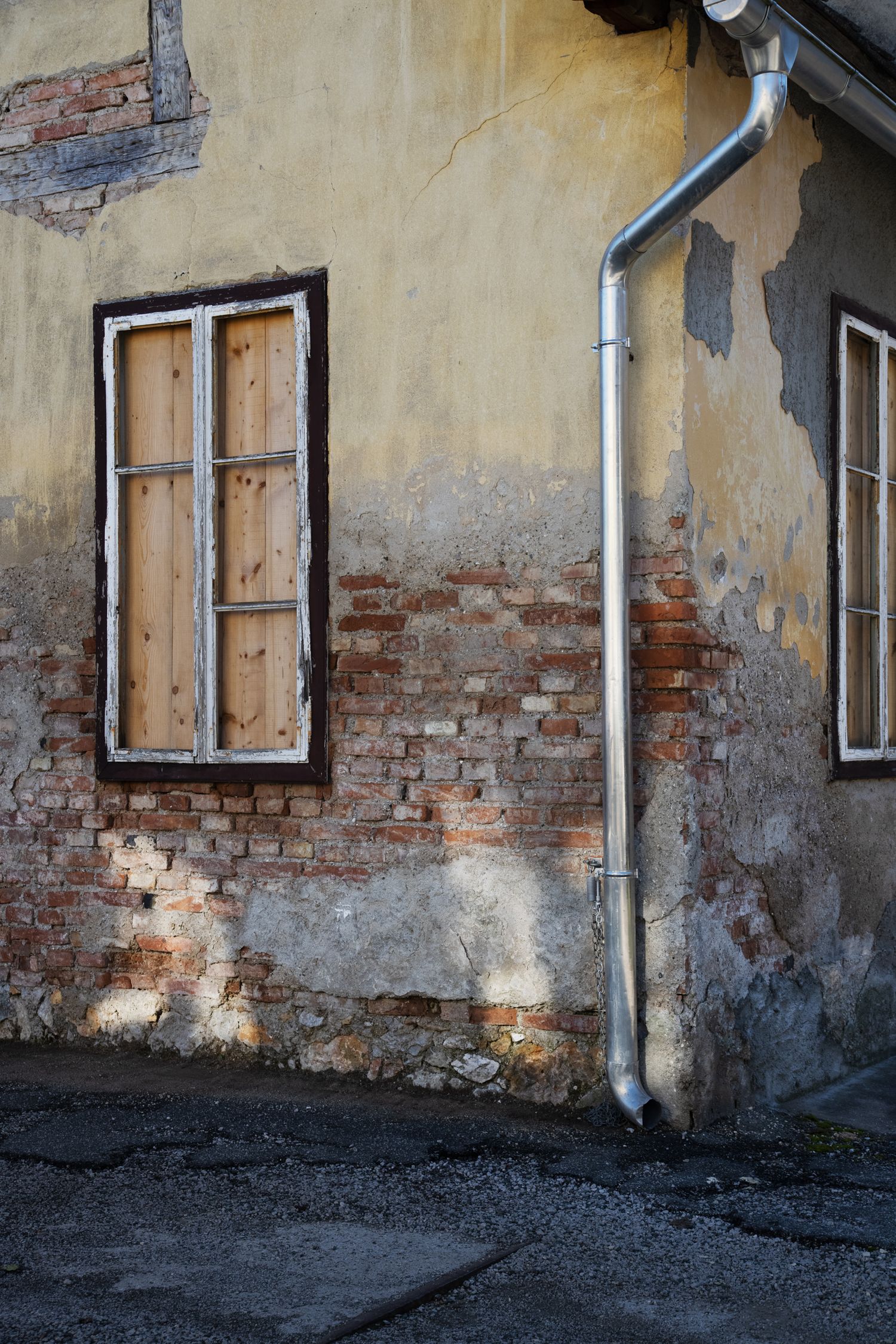 Weathered building exterior with boarded window, crumbling brick wall, peeling plaster, and metal drainpipe in natural light.