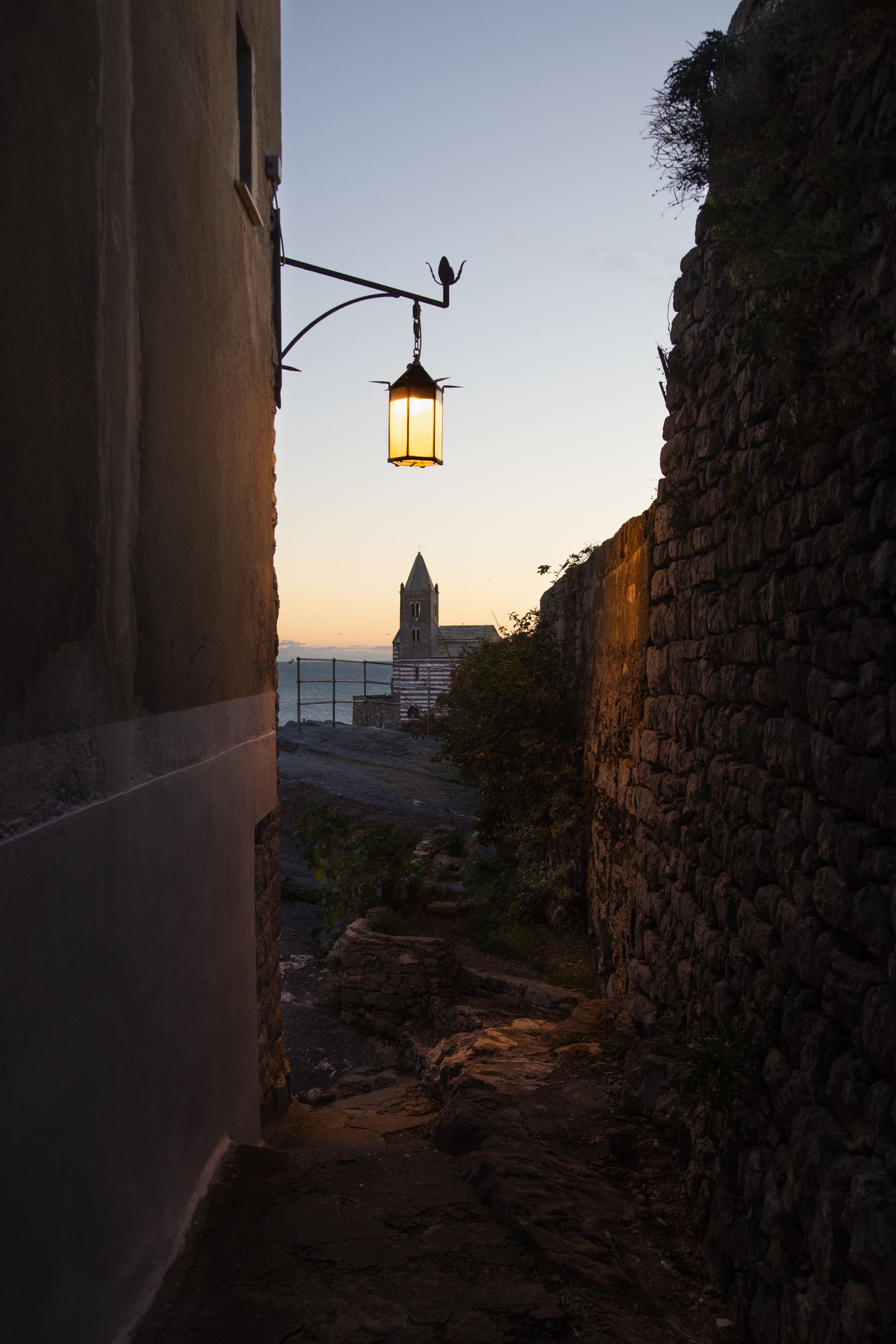 Narrow stone alley at dusk with a glowing vintage lantern and church tower view.