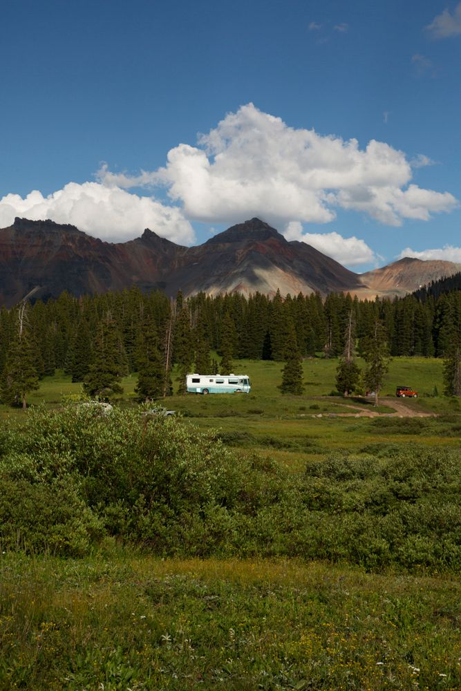 Mountain landscape with evergreen forest and meadow featuring a small camper in the distance.