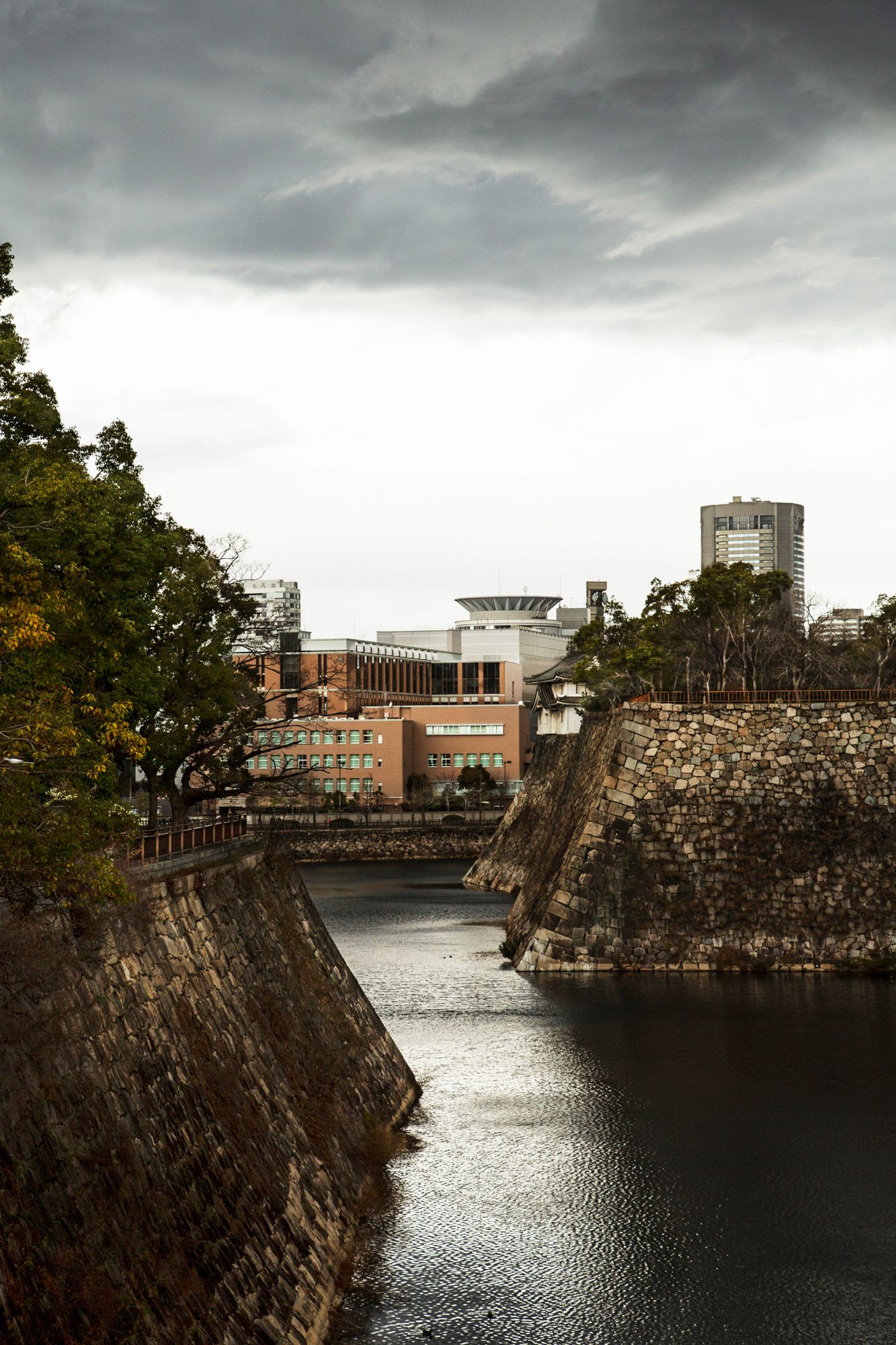 Dramatic storm clouds over a moat surrounding stone fortress walls in an urban setting.