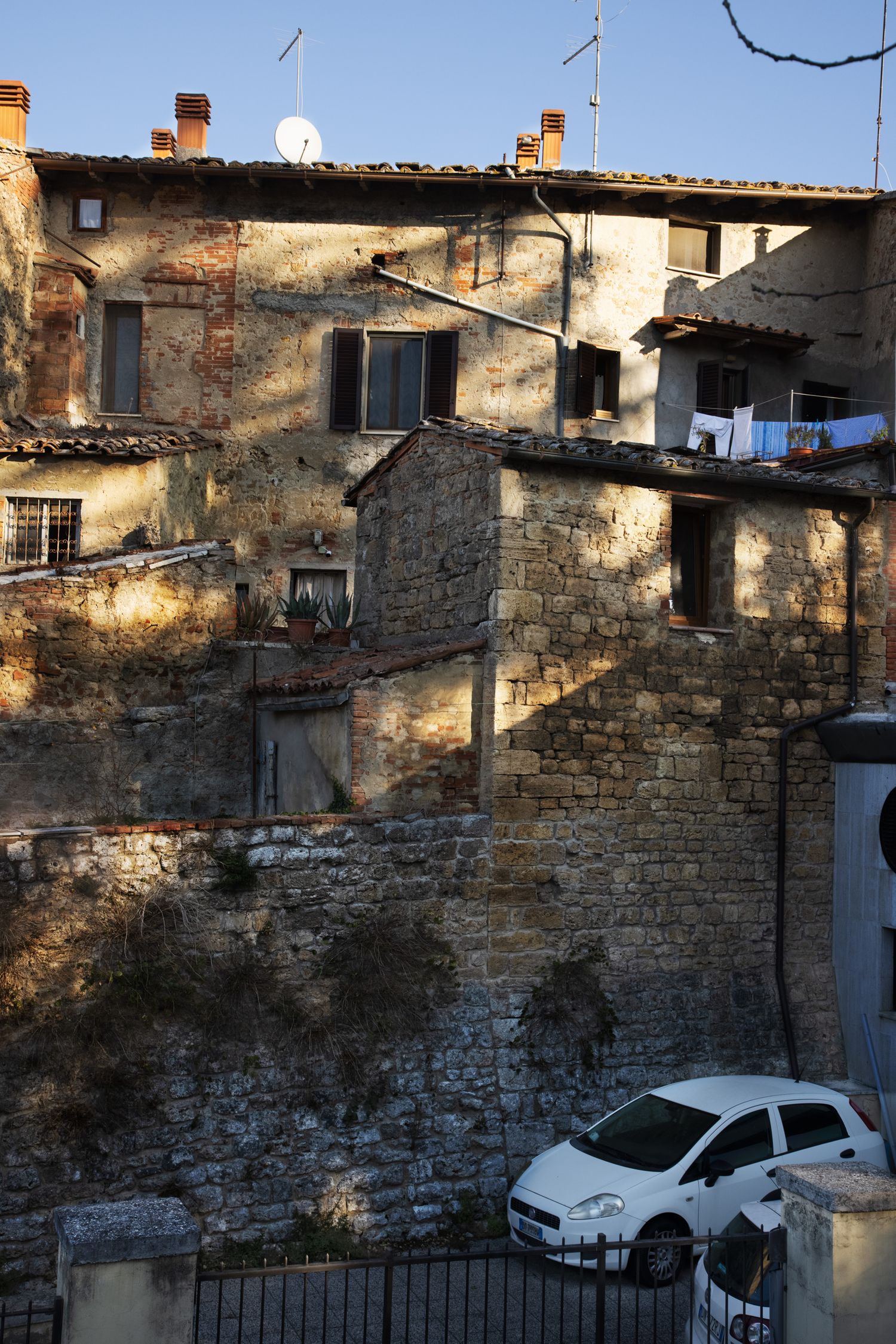 Ancient stone buildings in a historic Italian hilltop village with a white Fiat parked below sun-drenched medieval walls.