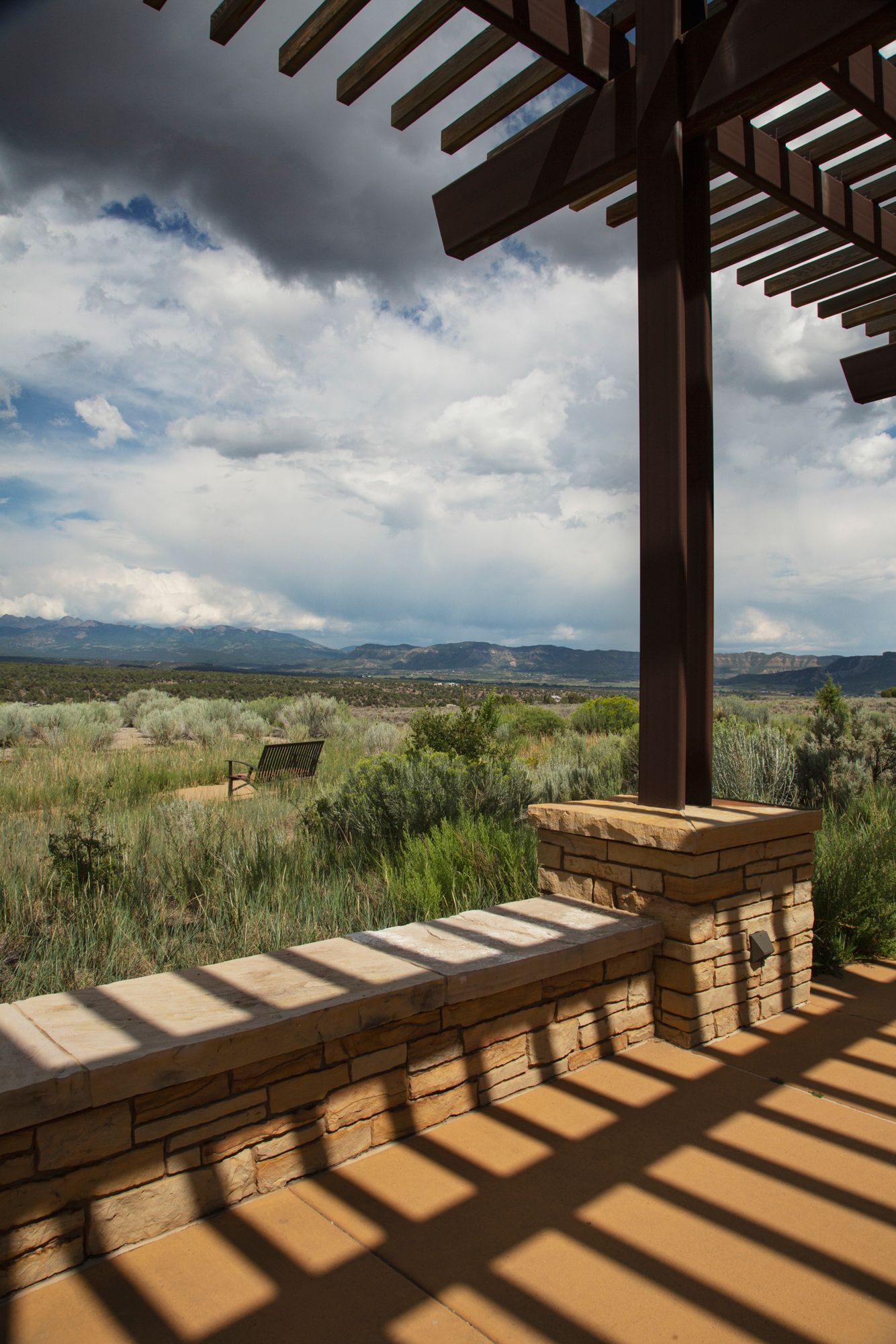 A covered patio with wooden beams casts dramatic shadows while overlooking a scenic mountain landscape view.