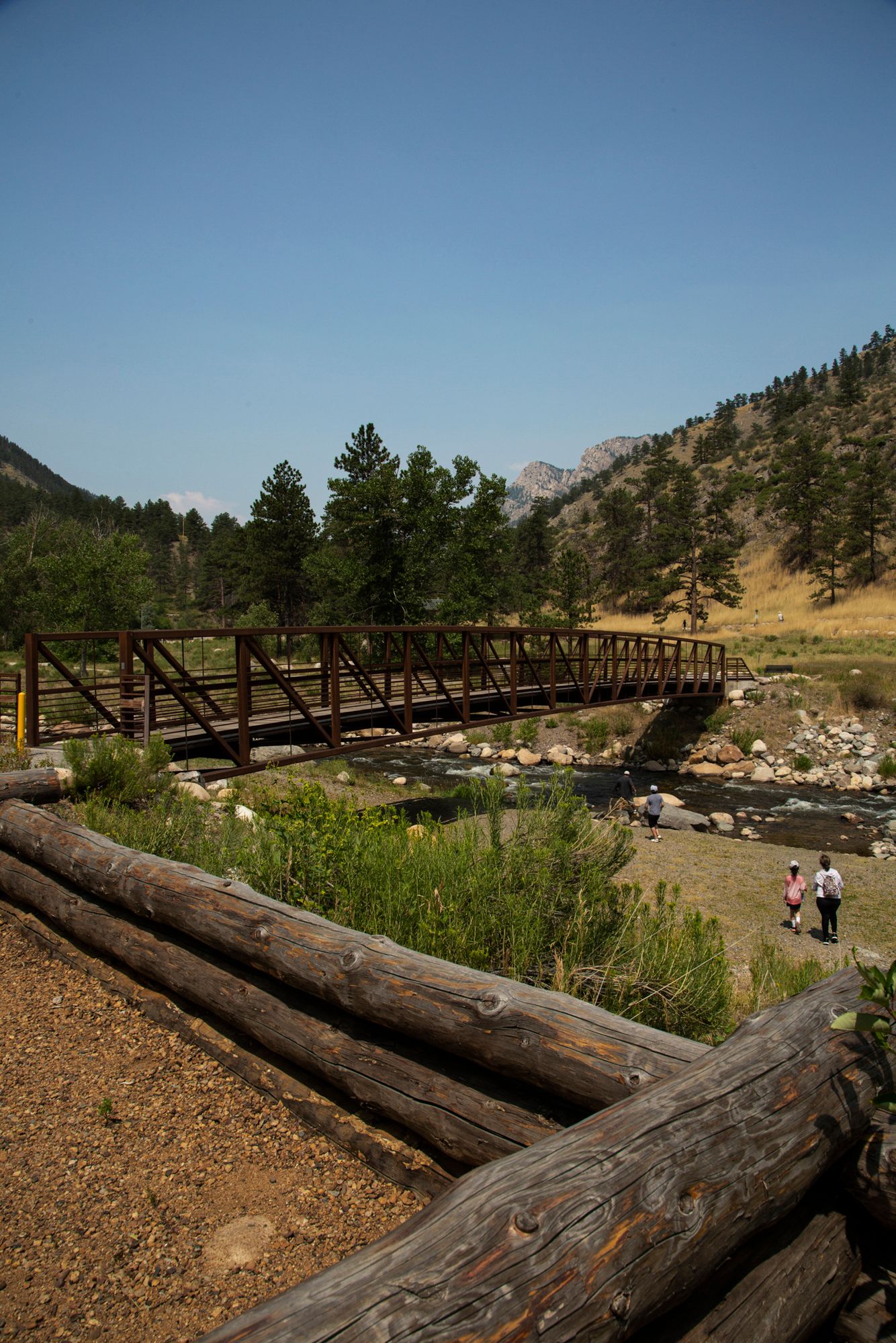 A rustic wooden bridge spans across a scenic mountain landscape with hikers walking along the trail on a sunny day.