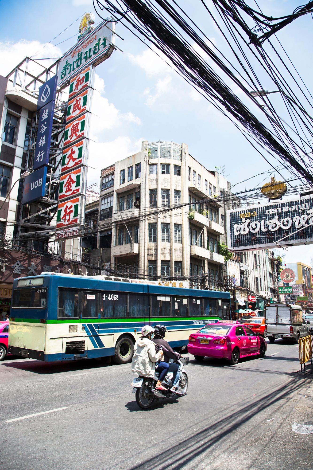 Busy Bangkok street with signs, electrical wires, public bus, and typical urban traffic.