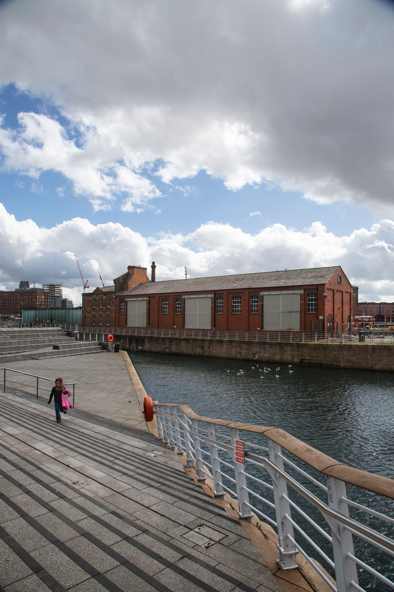 Wooden boardwalk with metal railings extends along an industrial waterfront with brick buildings.