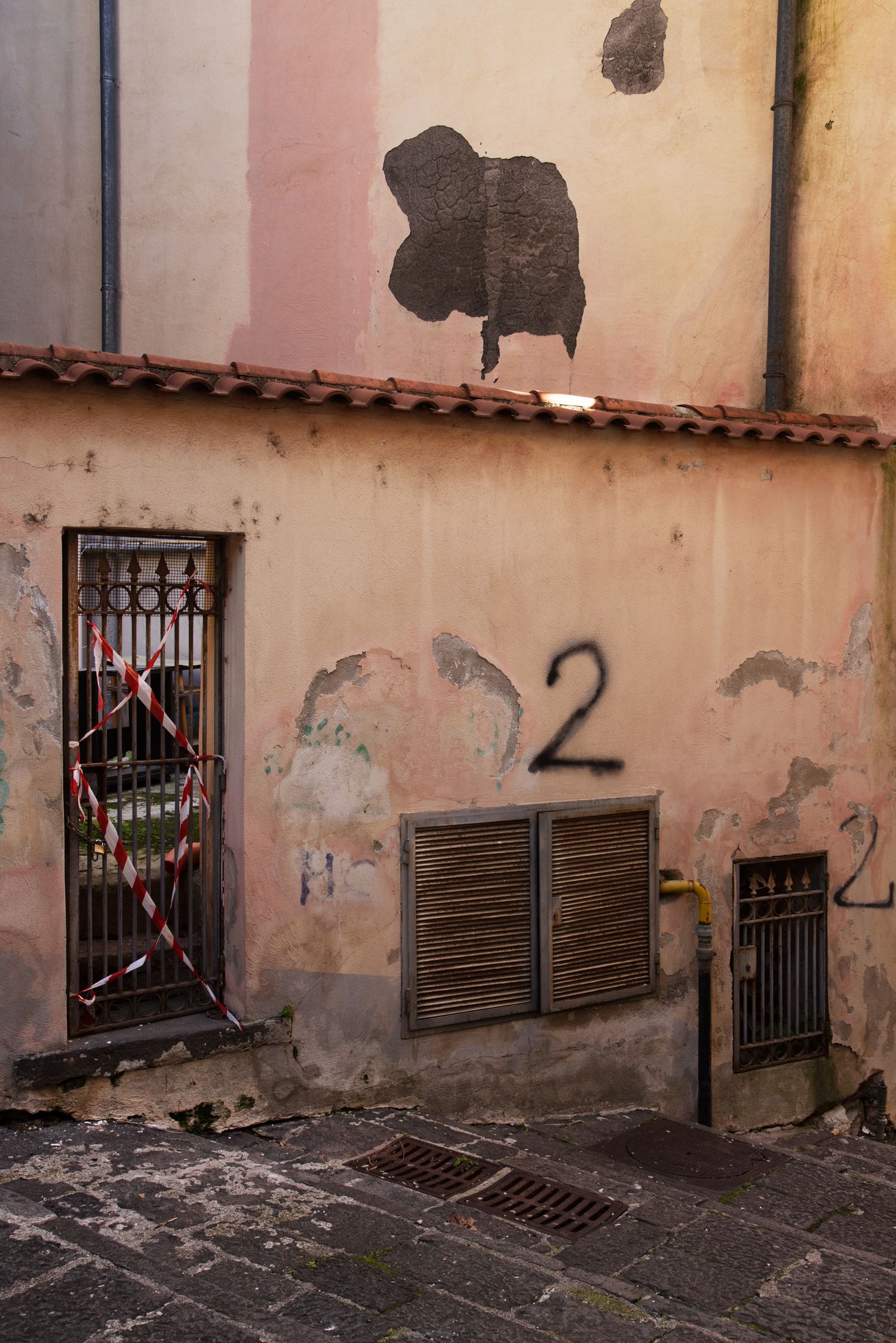 Weathered building facade with peeling paint, barred window with caution tape, wooden shutters, and graffiti number 2.