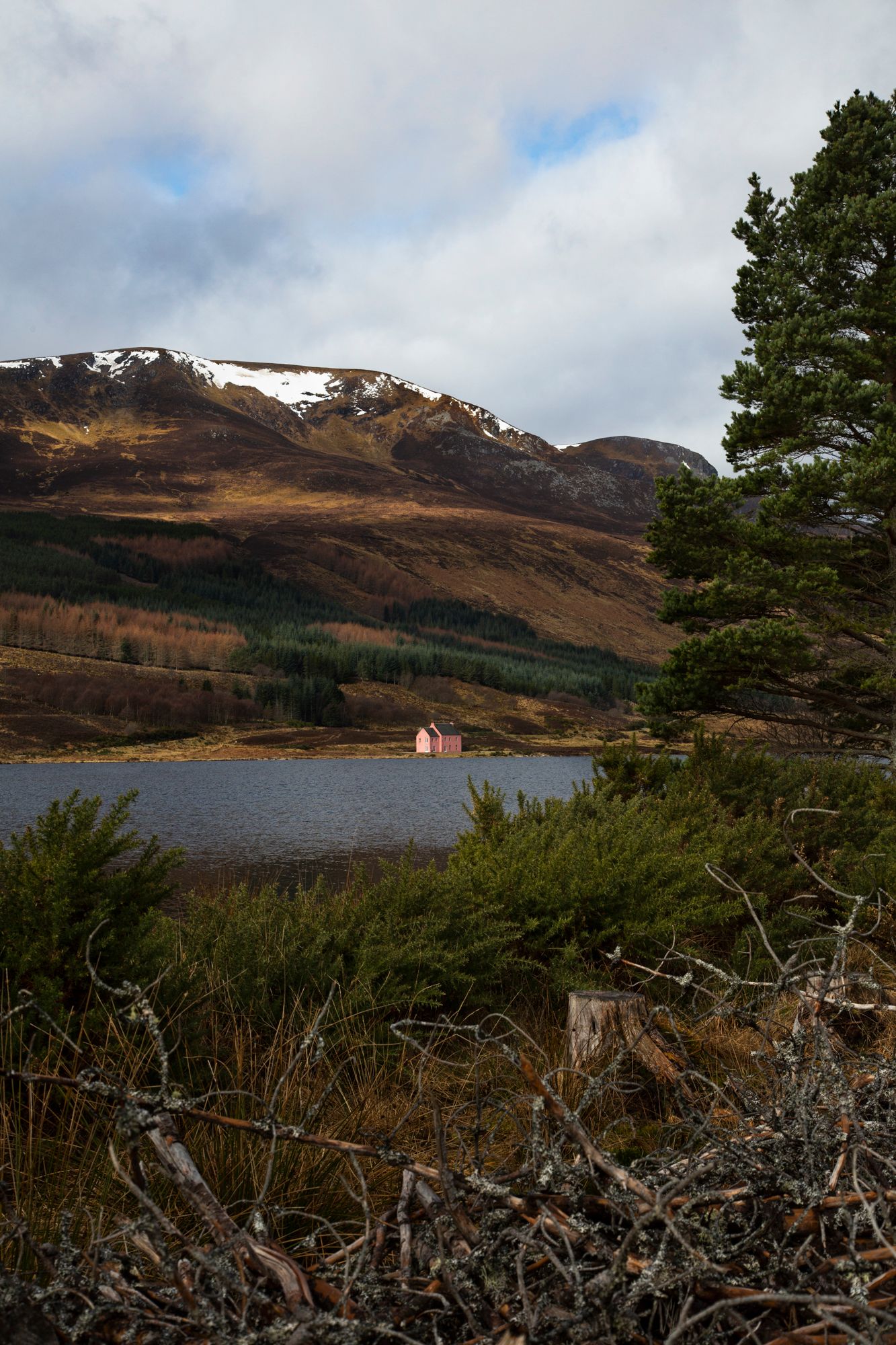Scenic Scottish Highland landscape with a small cottage nestled by a loch surrounded by rugged mountains and winter foliage.