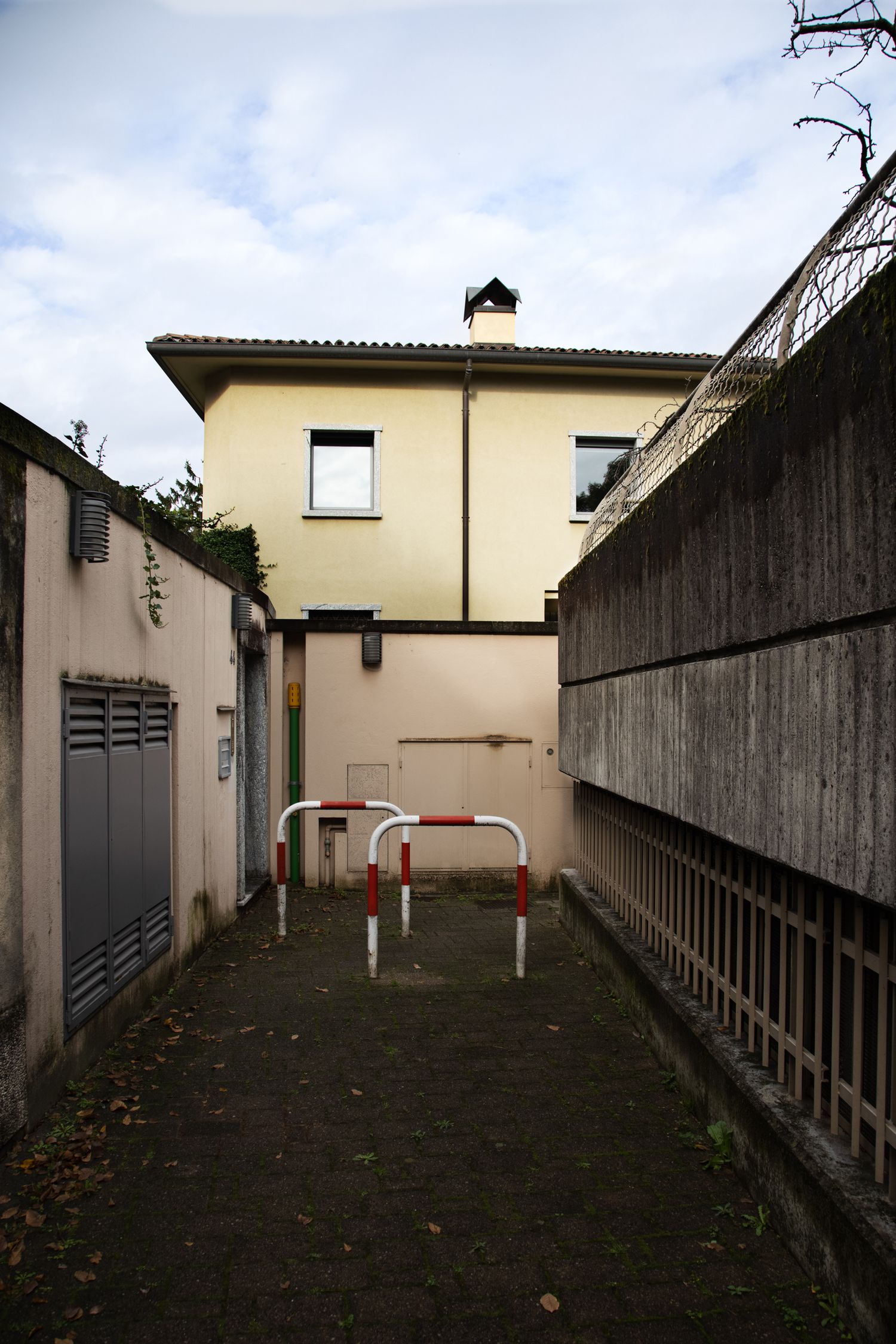 Narrow alleyway leading to a yellow two-story house with red and white barriers on a cloudy day.