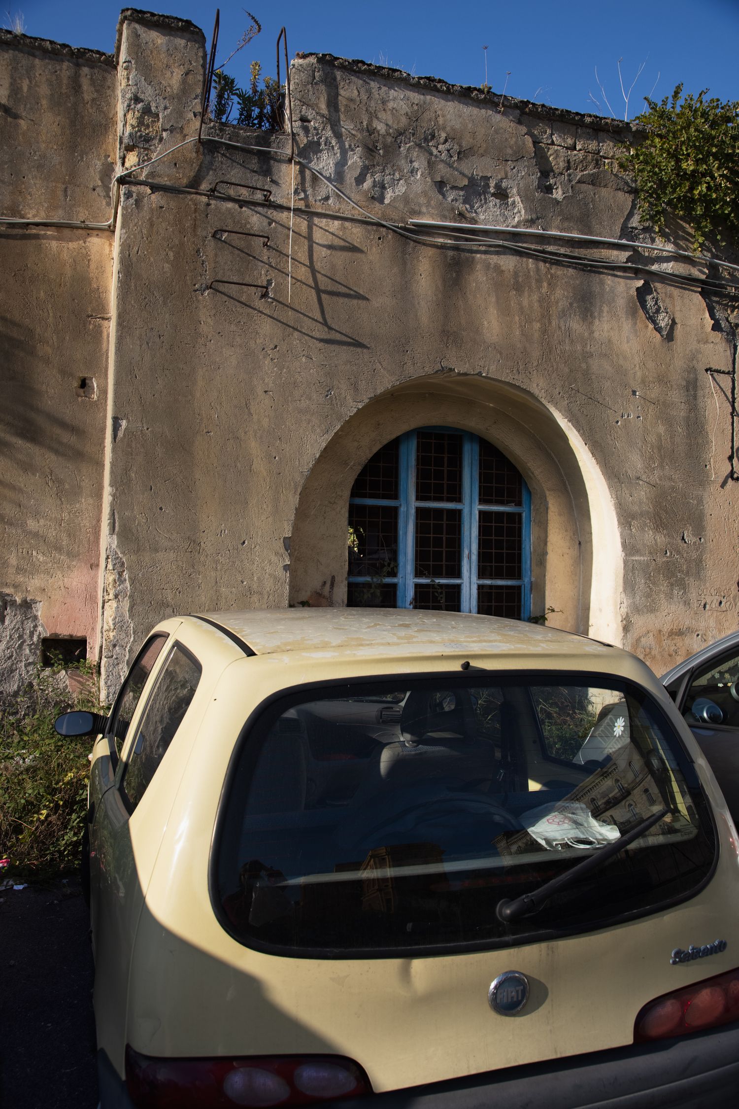A yellow compact car parked in front of a weathered stone building with a blue arched window in warm sunlight.