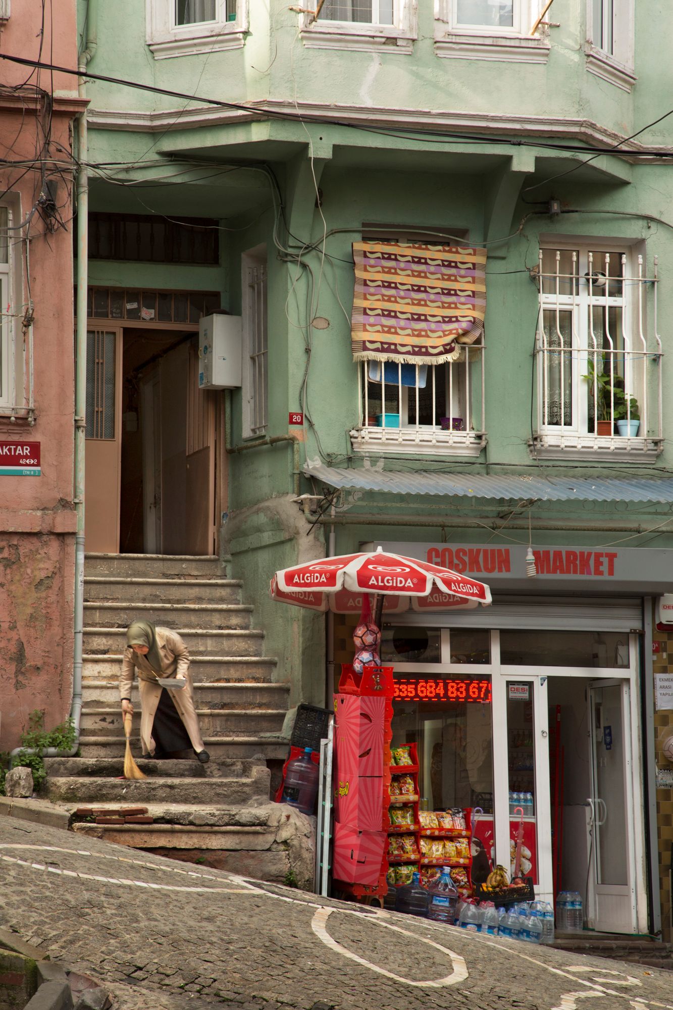 Corner market with red umbrella and stone steps leading to mint green building facade.