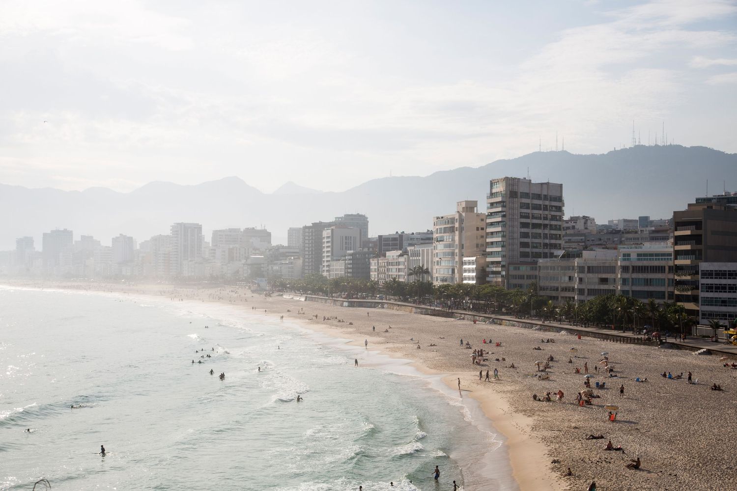 Panoramic view of a crowded beach with high-rise buildings and mountains in the background.