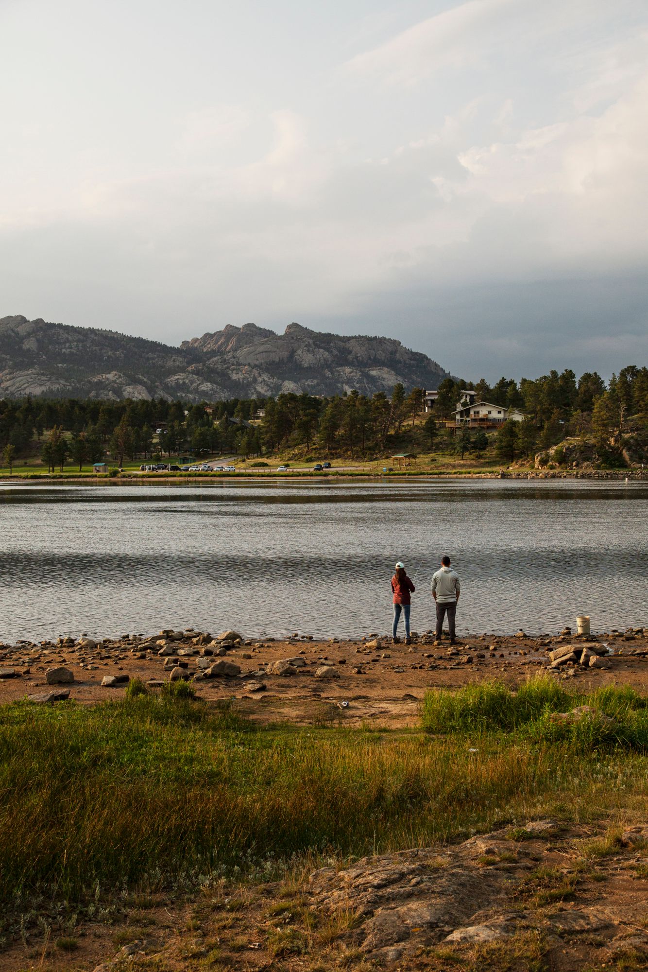 Two people stand at the rocky shore of a mountain lake during golden hour with rugged peaks and trees in the background.