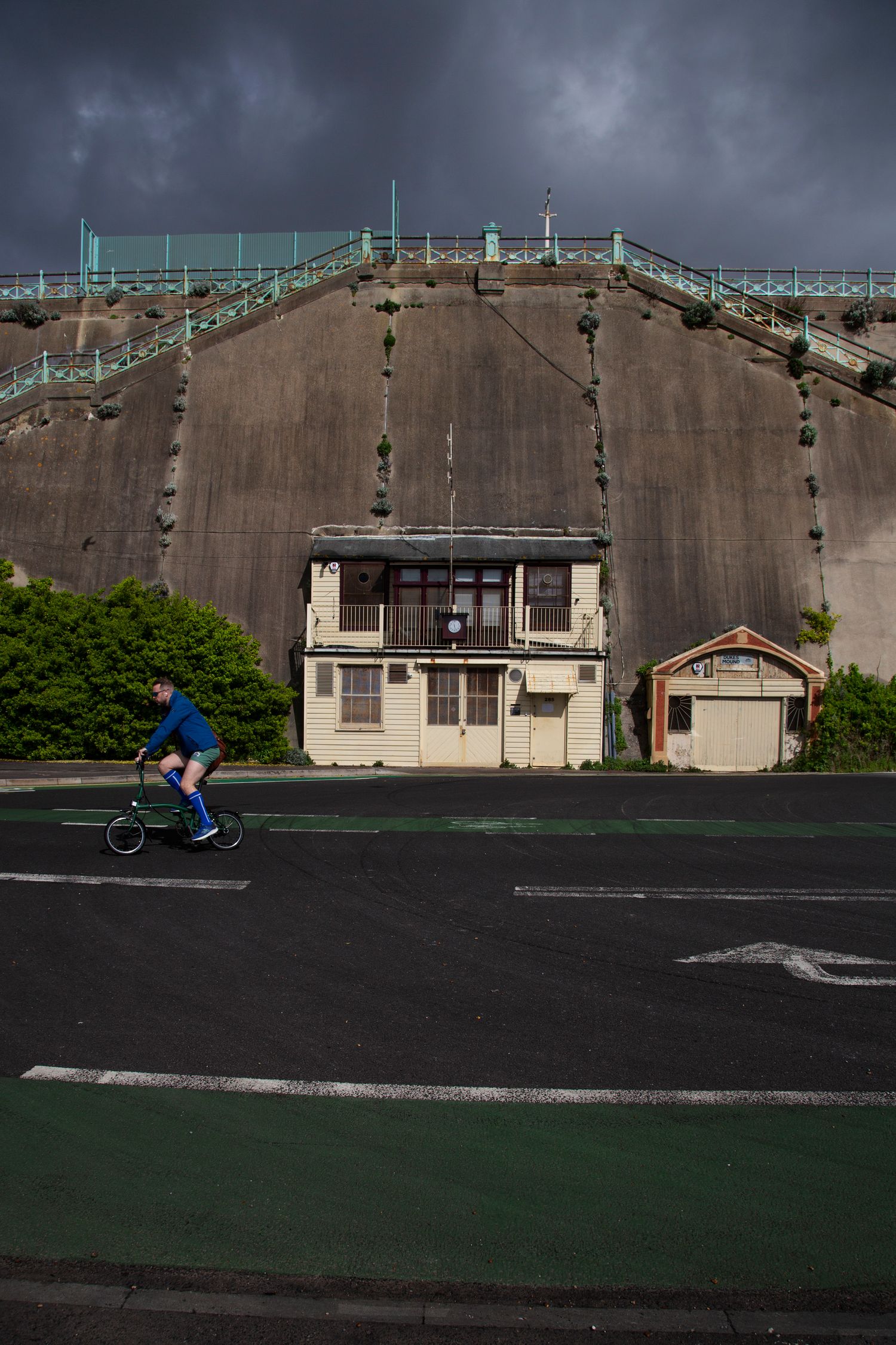 Cyclist passes large concrete structure with metal scaffolding under stormy skies.