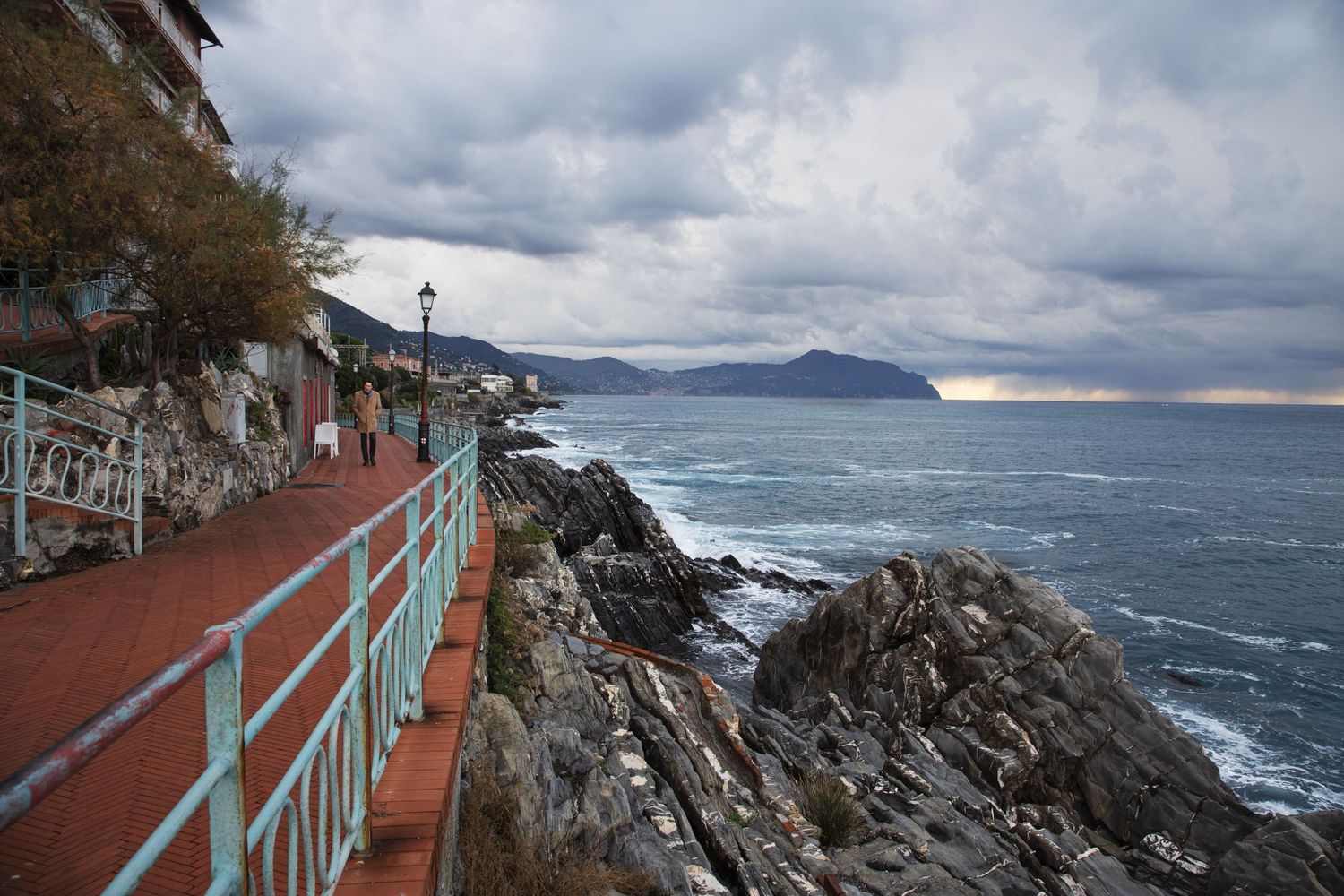 Scenic coastal walkway with teal railing along rocky shoreline under dramatic cloudy skies in Liguria, Italy.