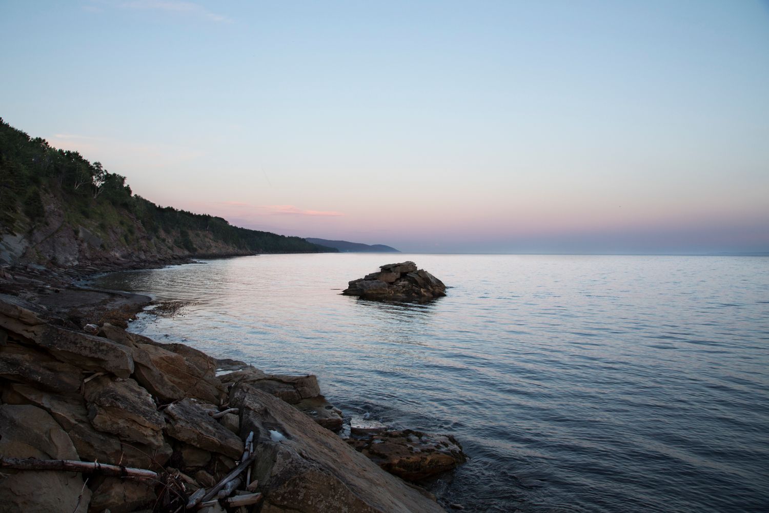 Serene coastal sunset view with rocky cliffs meeting calm ocean waters in soft pink light.