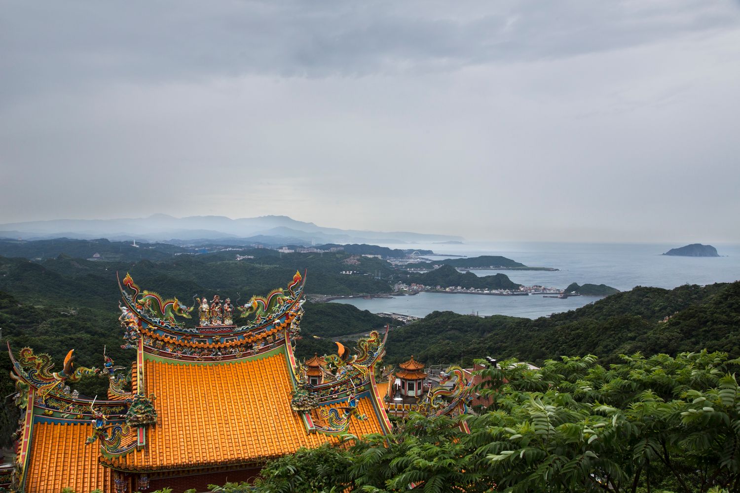 Temple with ornate orange roof offers sweeping views of mountainous coastal landscape.